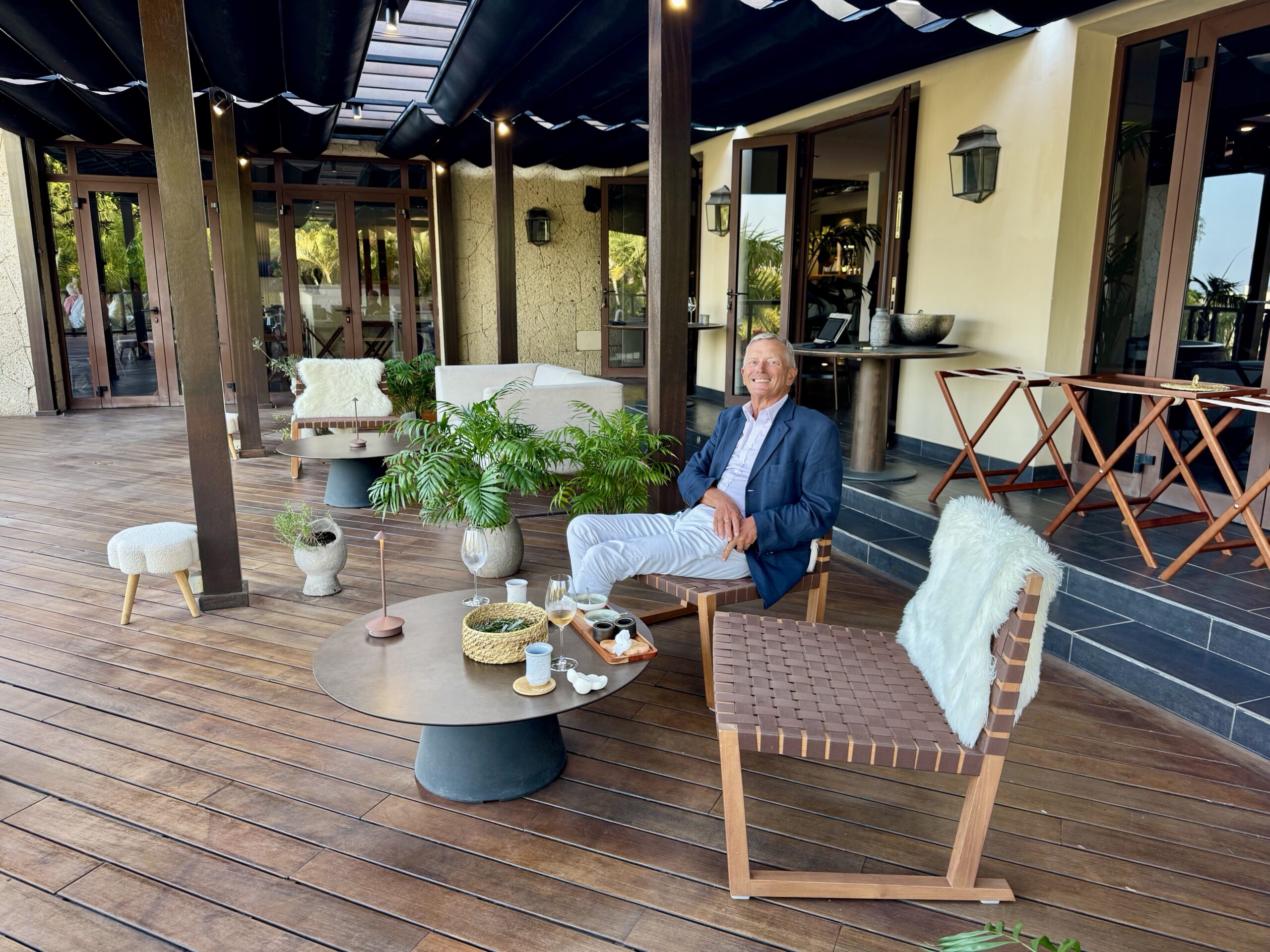 Smiling man in a blue blazer sits on a wicker chair on a wooden patio with tables, plants, and glass doors in the background.