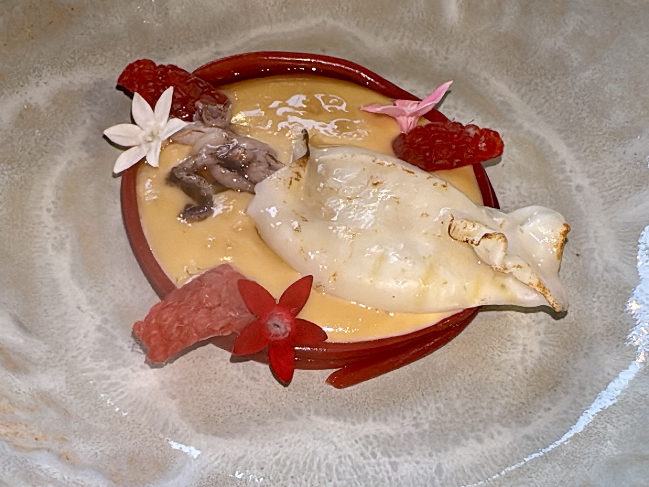 Plate with a dessert, yellow sauce, raspberries, and edible flowers as garnish (white flower and pink petals).