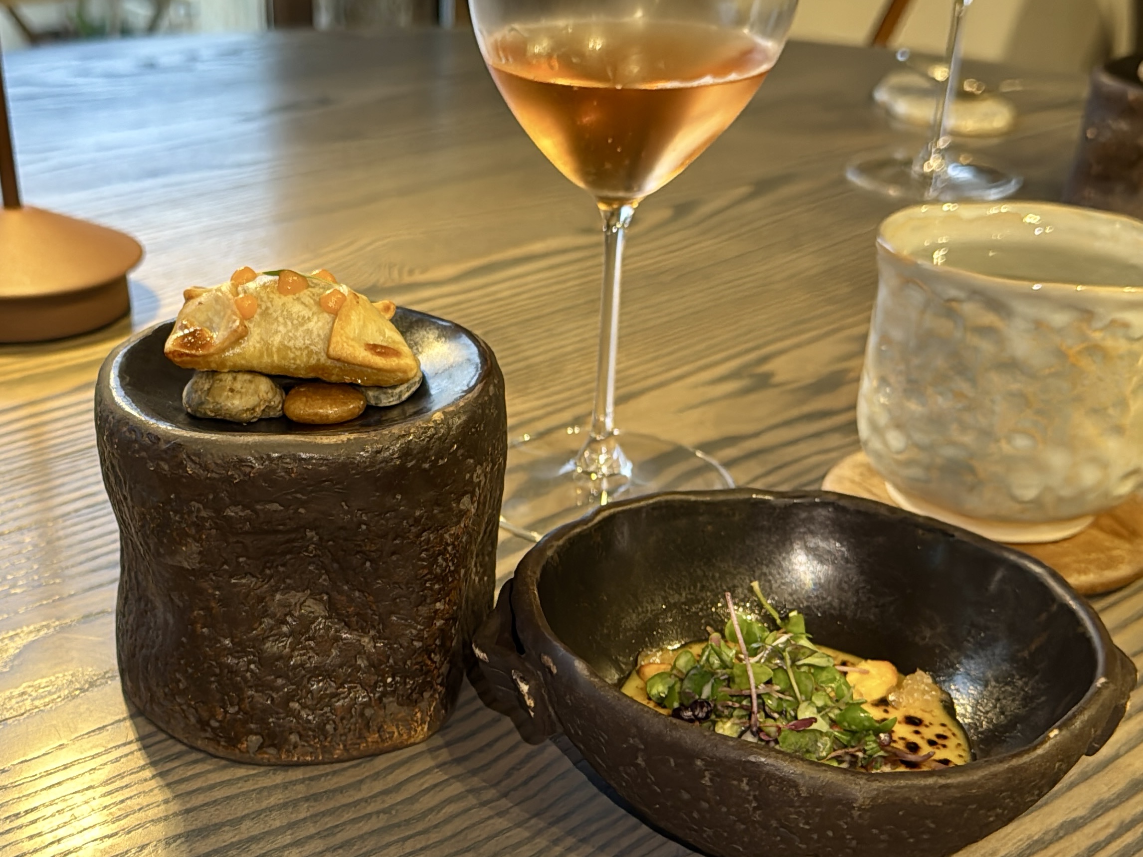 Small pastry canapé atop a black stone bowl on a wooden table, with a glass of wine beside.