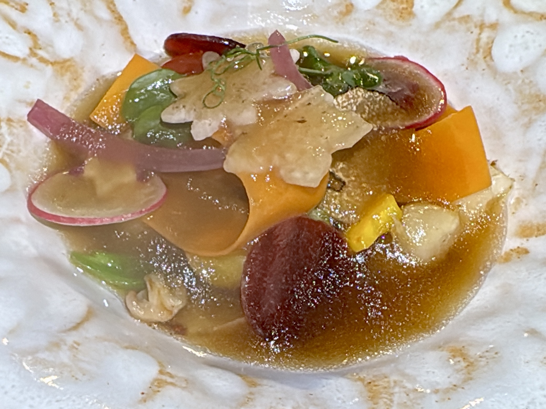 Close-up of a colorful, bite-sized flatbread topped with radish slices, orange pepper, greens, and a glossy sauce.