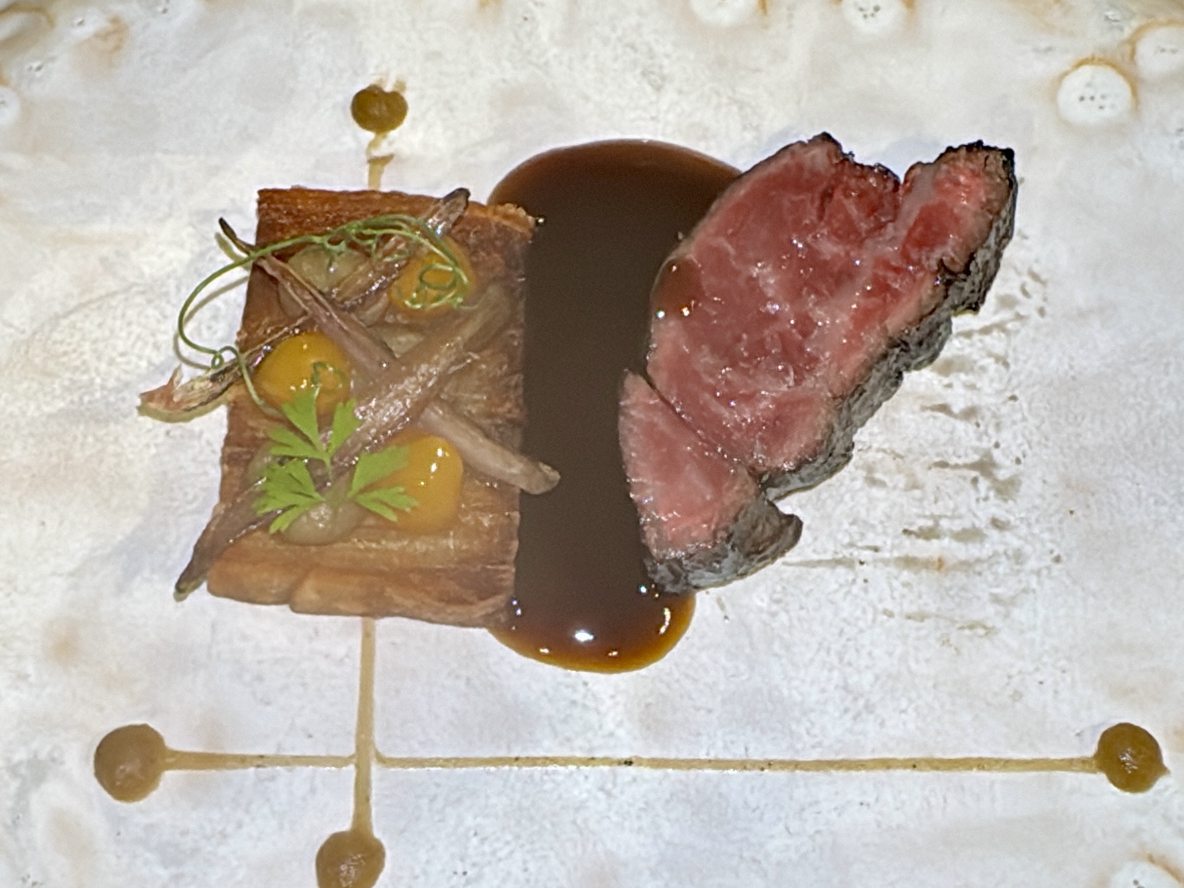 Two slices of beef with pink center, brown crust, drizzled sauce and a small herb garnish on a white plate.