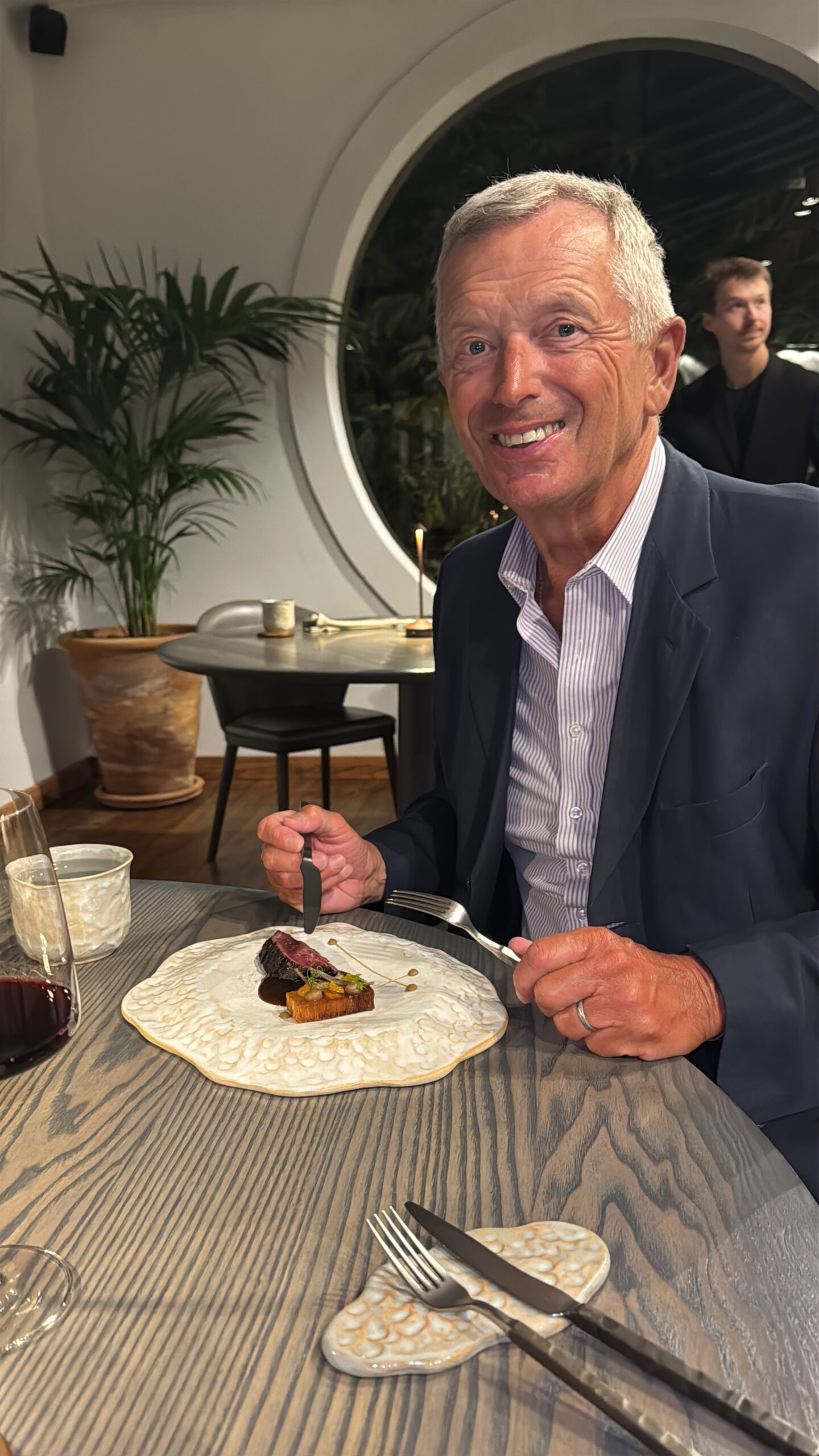Smiling older man in a navy blazer dining at a table with a plated steak and sauce, wine glass nearby.