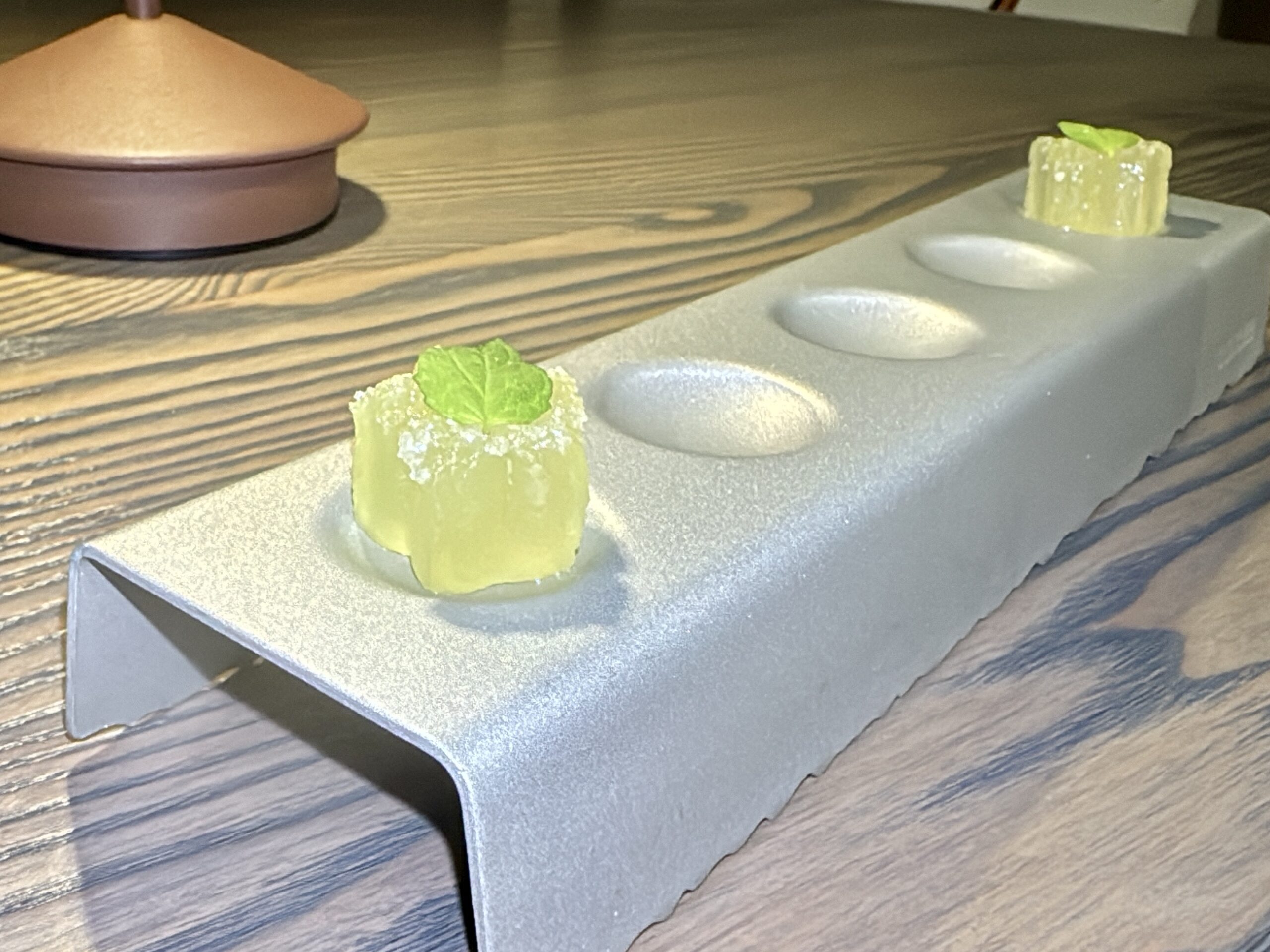 Three small yellow dessert cubes with mint leaves arranged on a silver tasting tray dusted with sugar, set on a wooden table.