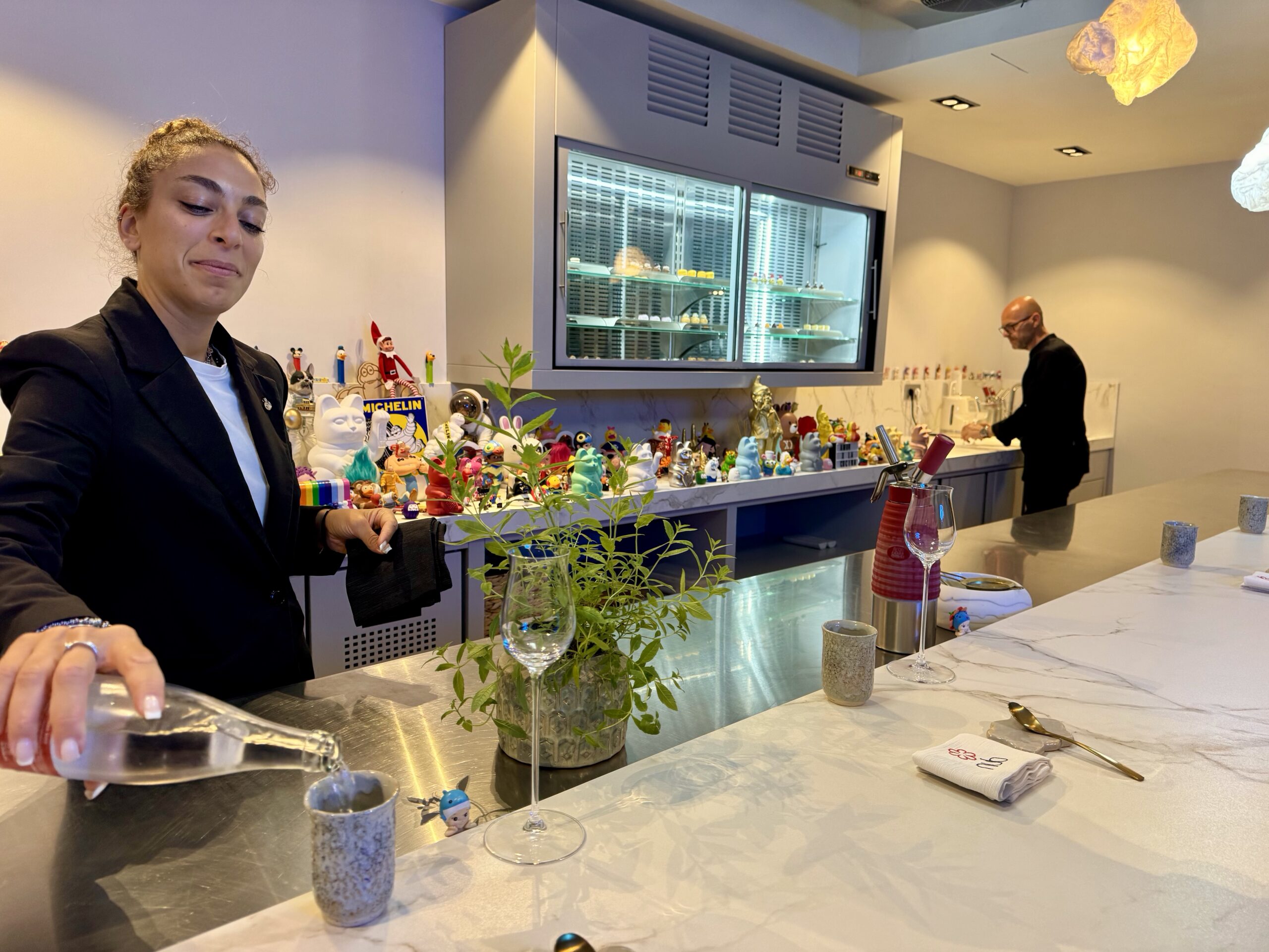 Female server in a black blazer pours a drink at a marble bar, with plants and colorful figurines along the counter; a man stands near a stainless cooler in the background.