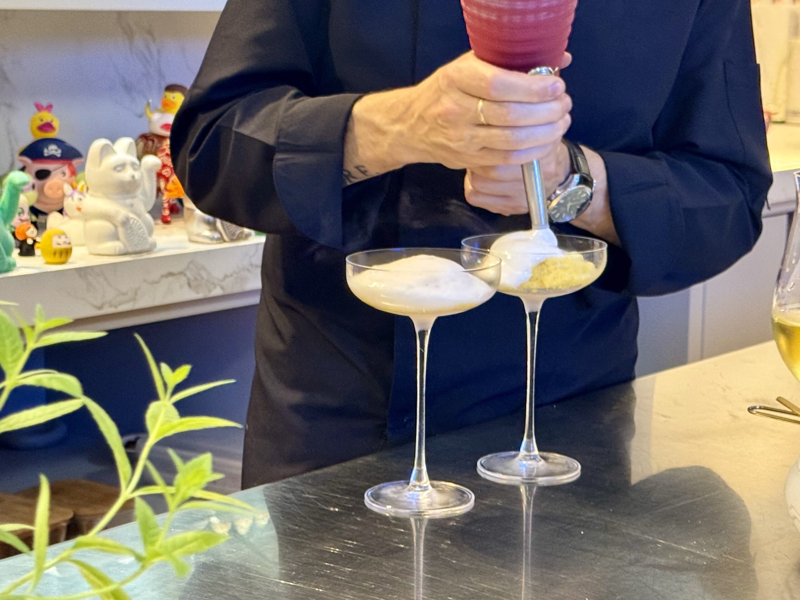 Person in a dark shirt scooping cream into two cocktail glasses at a bar, with colorful toy figurines on a shelf in the background