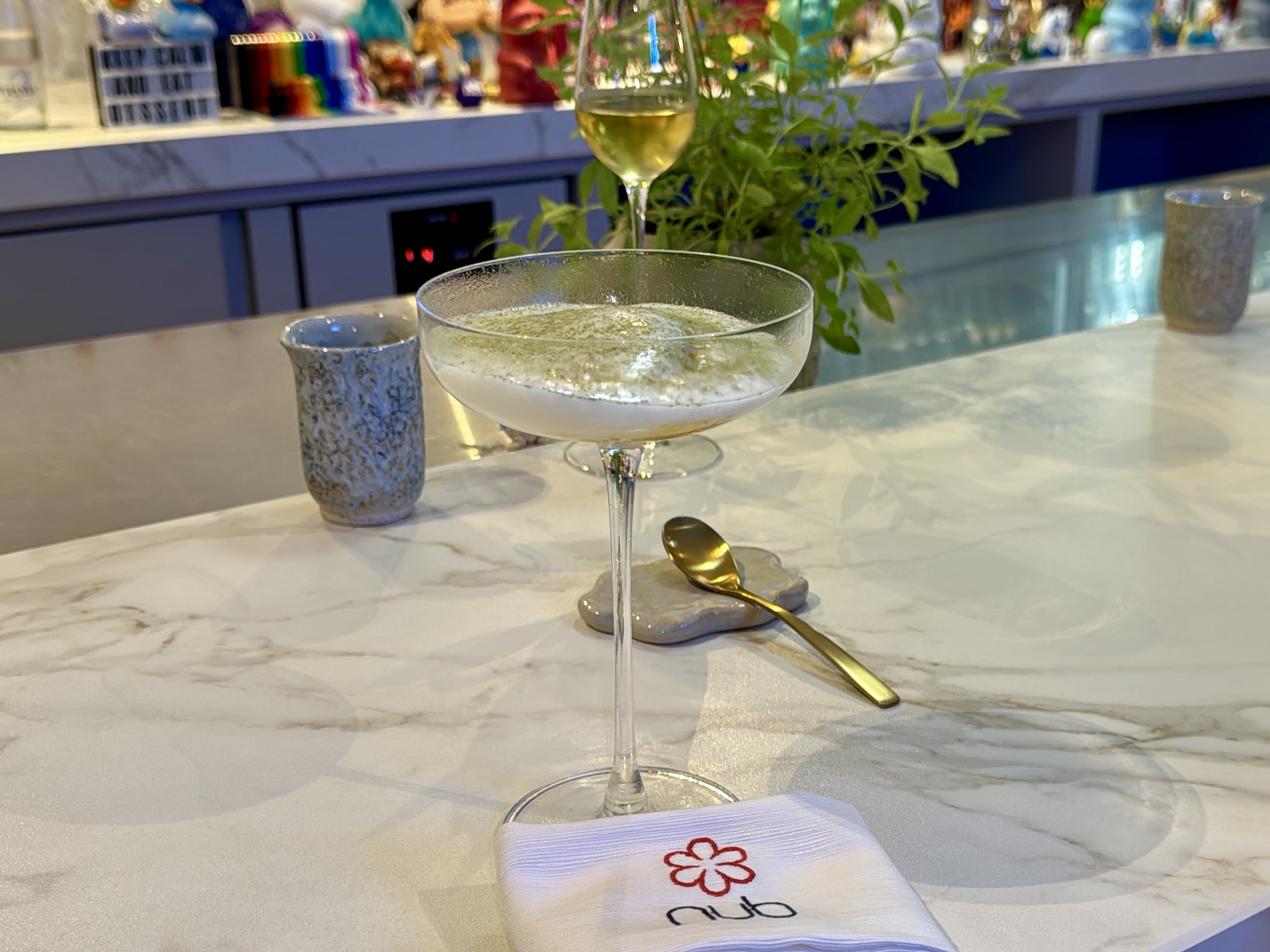 Close-up of a pale cocktail in a stemmed glass on a marble bar, with a napkin and golden spoon in the foreground and a plant in the background.