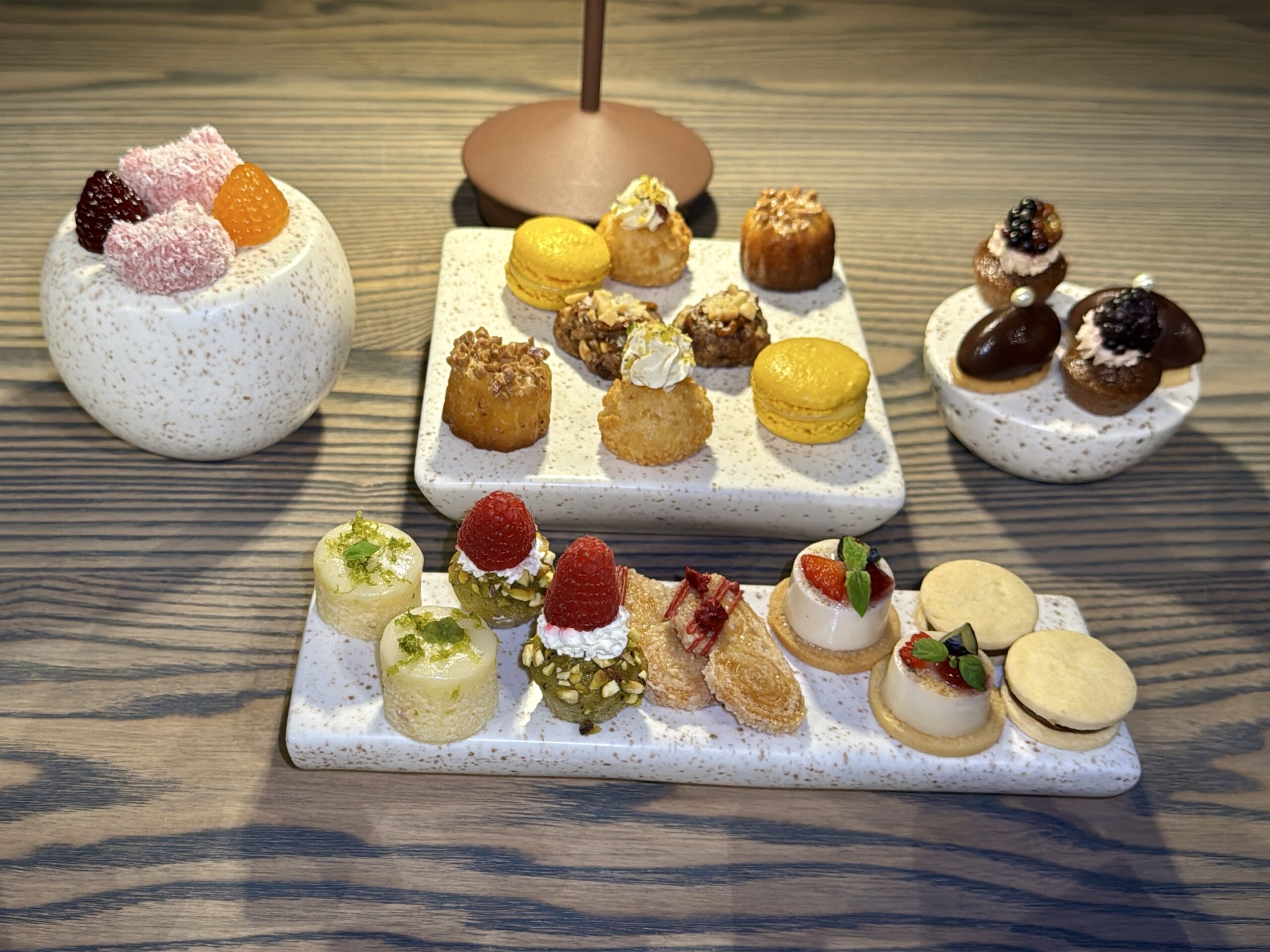Assorted bite-sized desserts on speckled white platters, including macarons, cream puffs, chocolate pastries, and berries on a wooden table.
