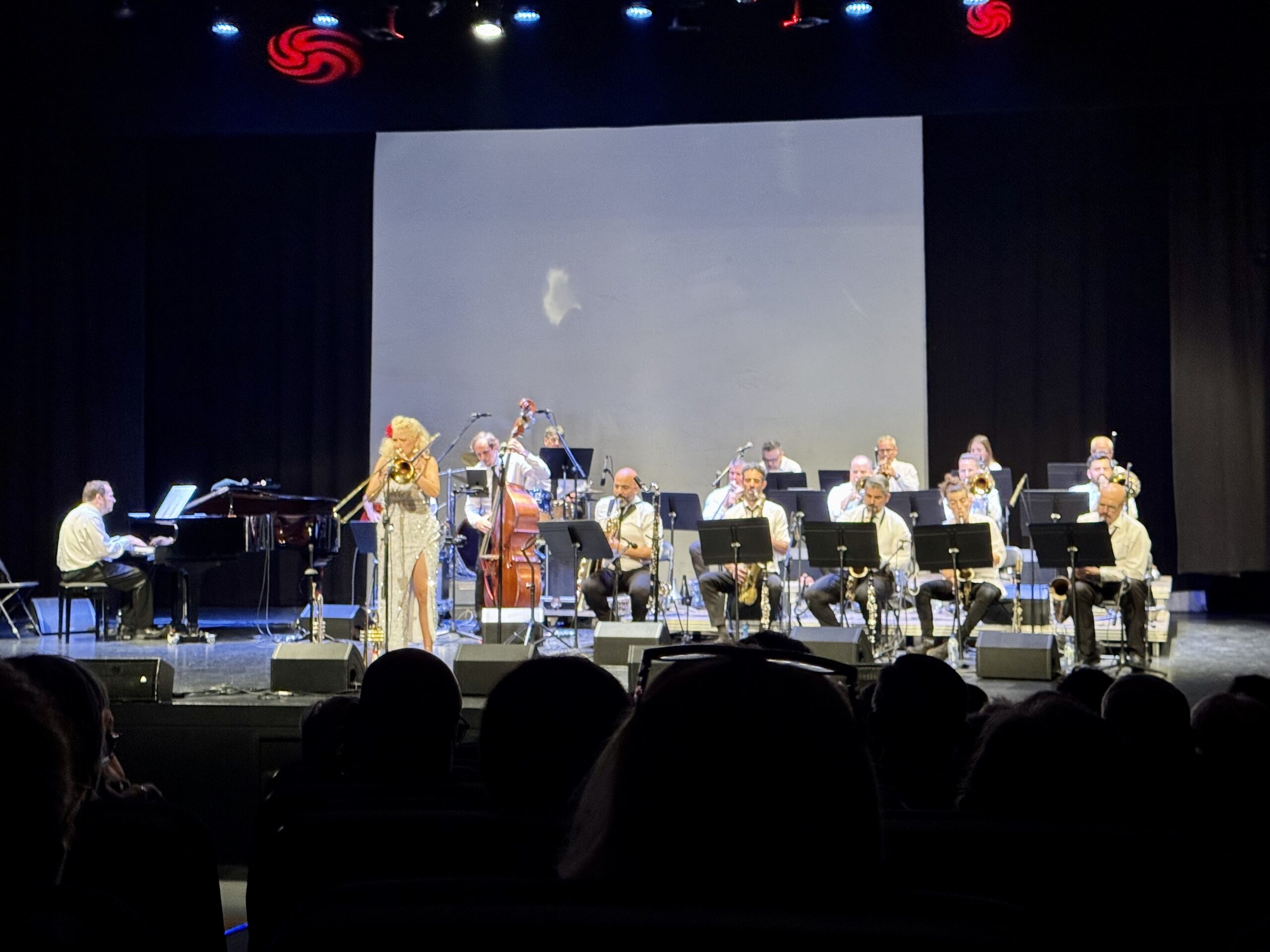 Wide view of a live concert: a brass band on stage with a female singer at center, pianist to the left, and musicians with sheet music stands behind her; audience silhouettes in the foreground under stage lights.