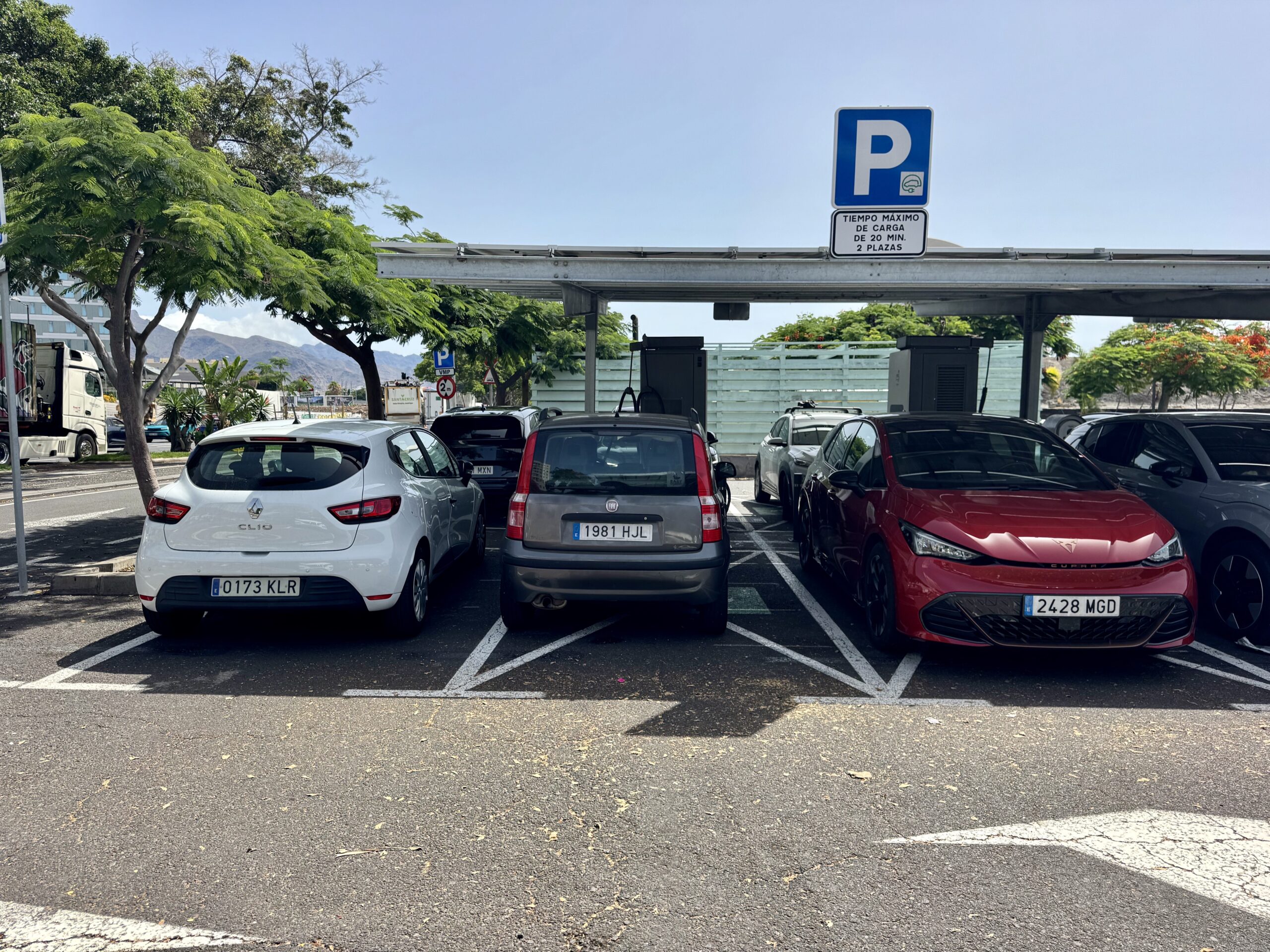 Parking lot filled with cars under a covered area, with a blue parking sign overhead and trees in the background.