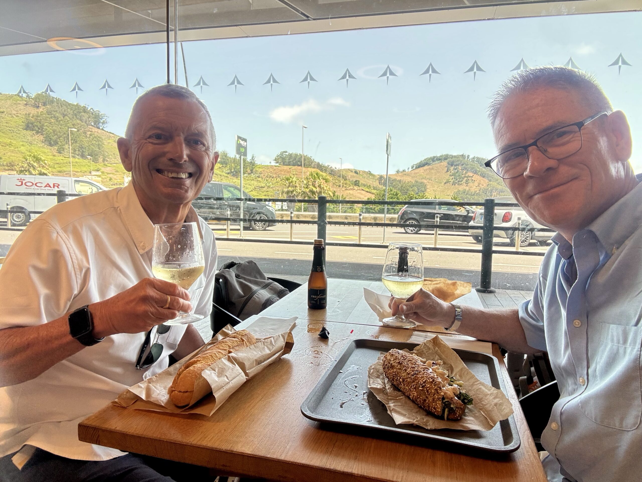Two men sit at an outdoor cafe, clinking wine glasses with baguettes on the table, smiling at the camera.