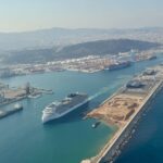 Aerial view of a busy harbor with a large white cruise ship docked at a port beside cranes and warehouses.