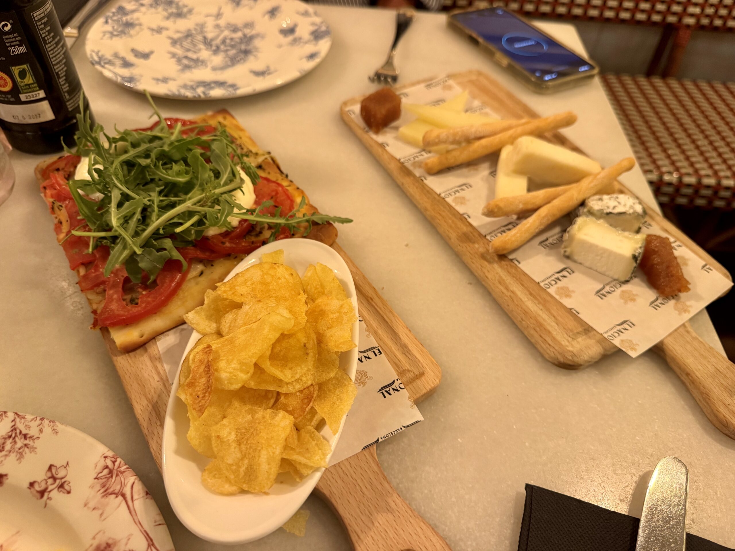 Wooden boards with bread, cheese, and breadsticks alongside a plate of arugula-topped flatbread and tortilla chips on a table.