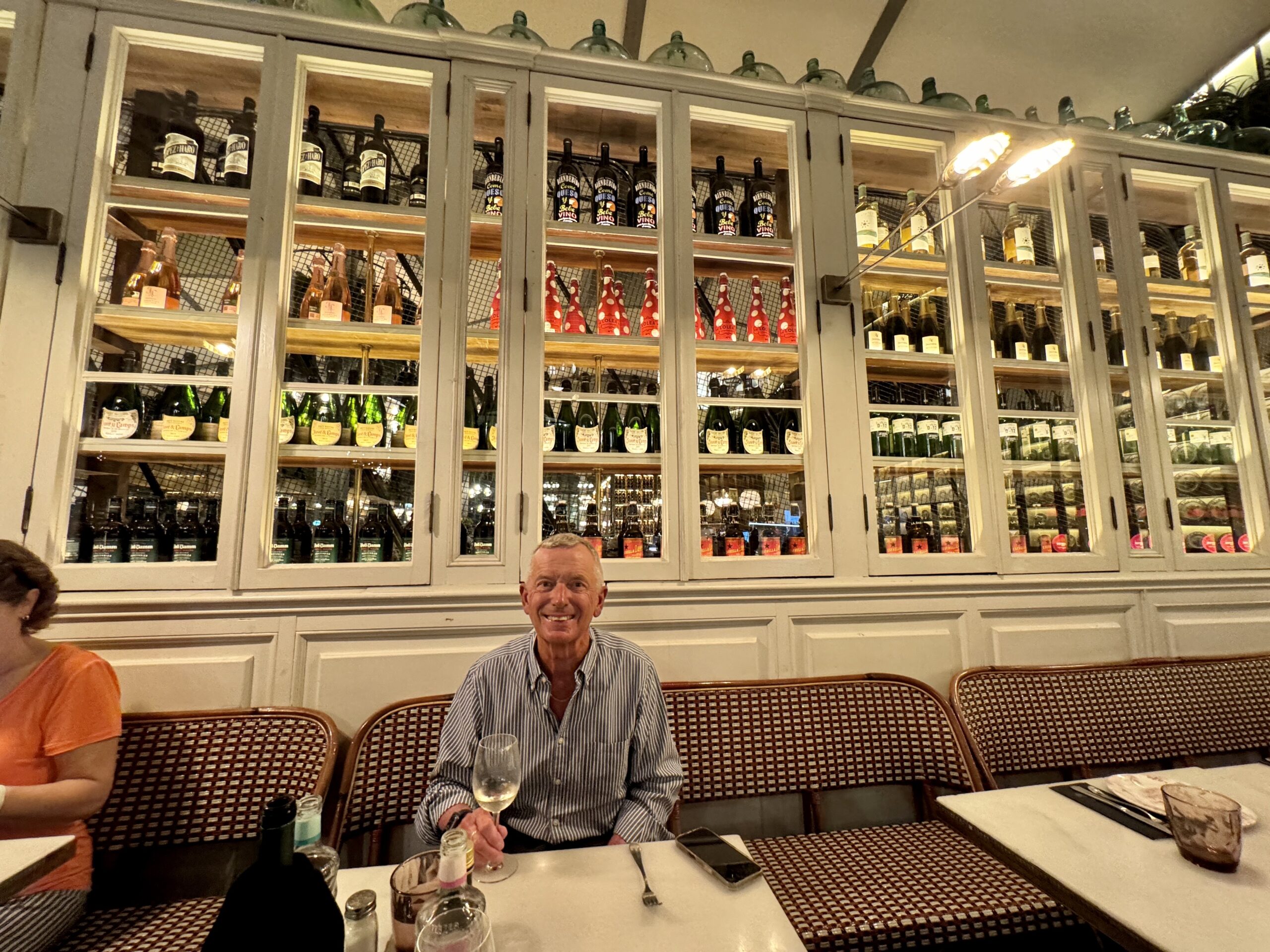 Smiling man in a striped shirt sits at a table in a wine-cluttered restaurant with glass-fronted shelves of bottles behind him.
