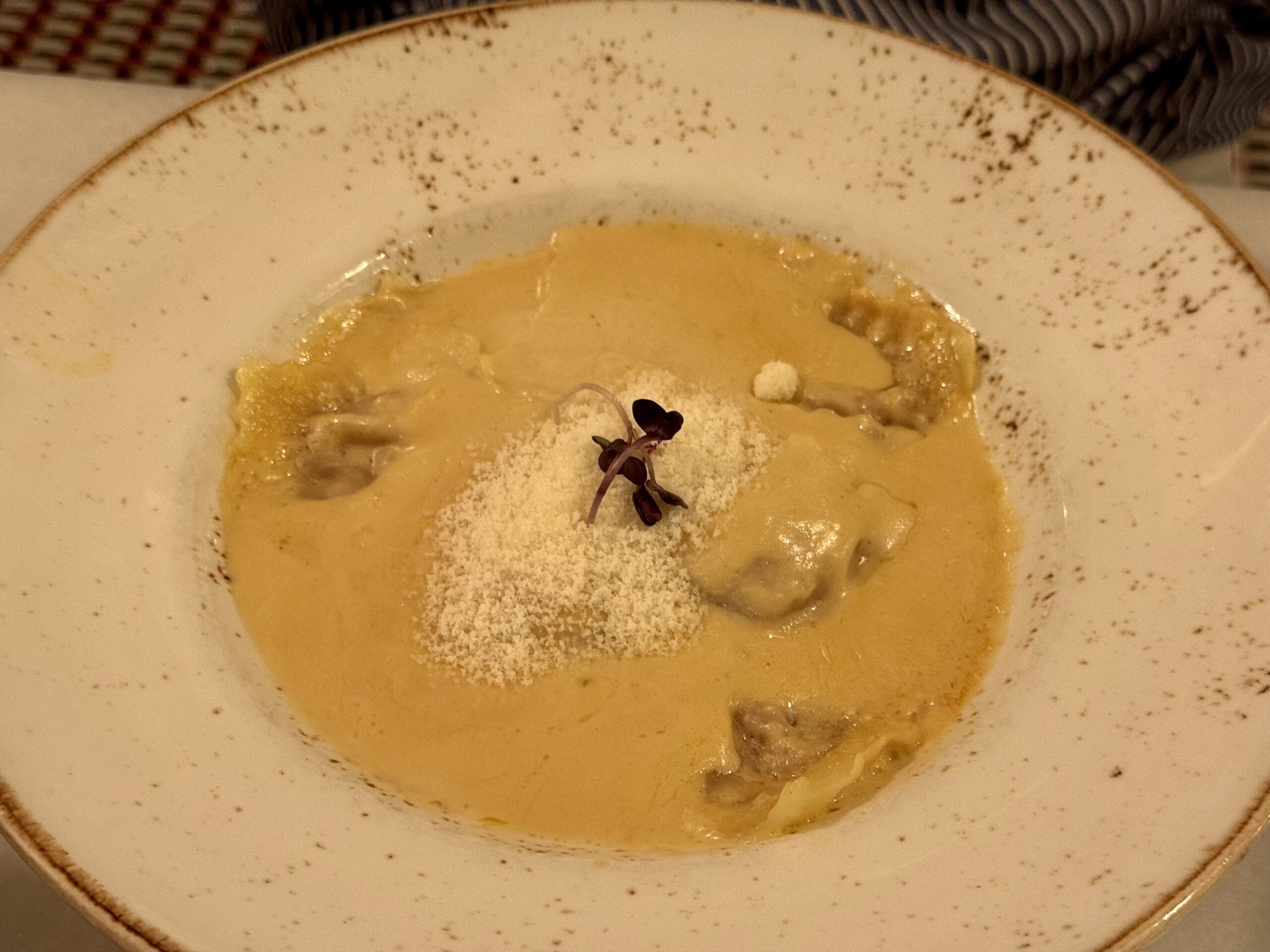 Pasta in a yellow sauce on a speckled white plate, garnished with a small green herb sprig and grated cheese.
