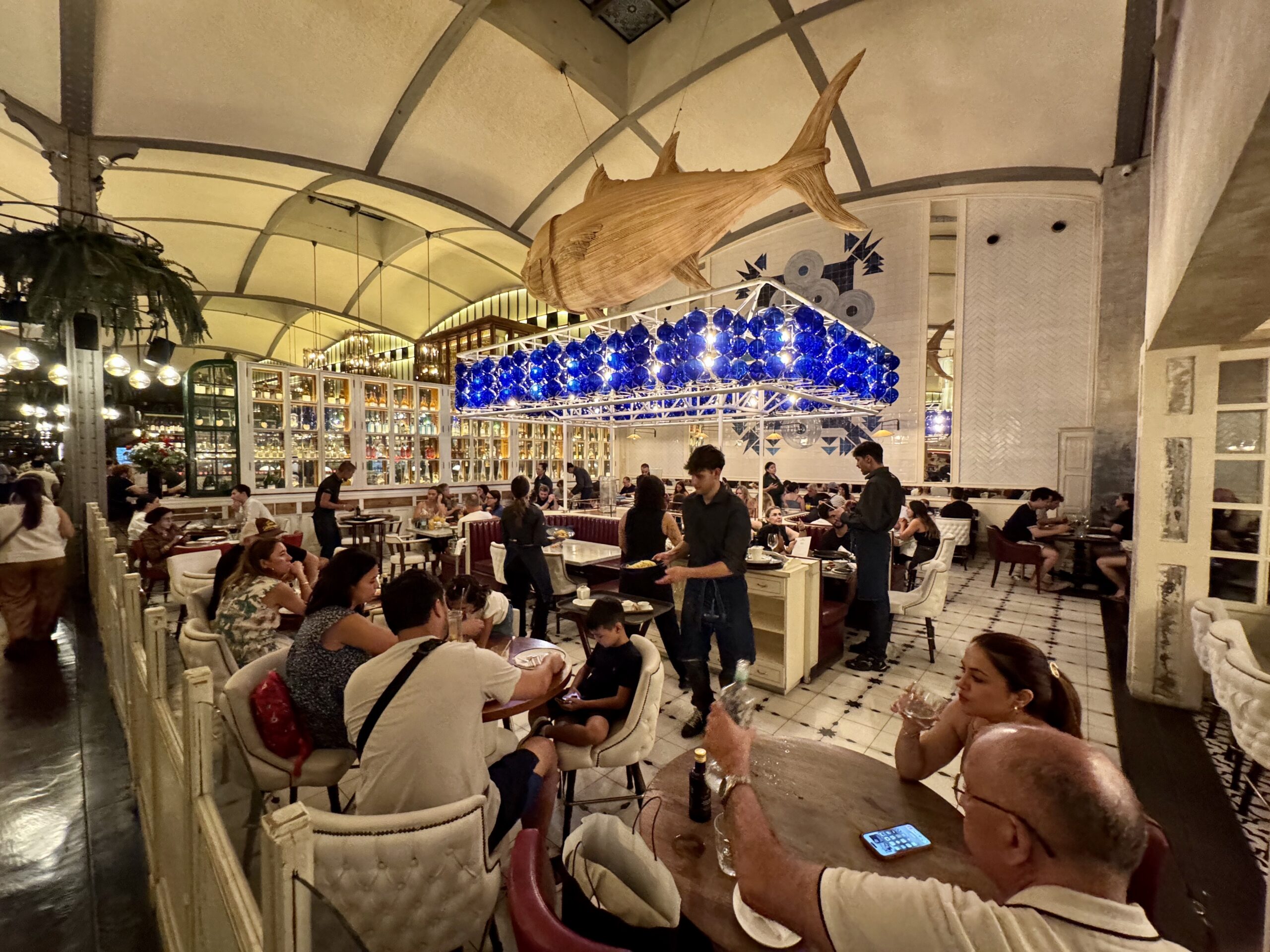 Inside a lively restaurant with a large wooden whale hanging from the ceiling and a blue glass sphere chandelier above diners.