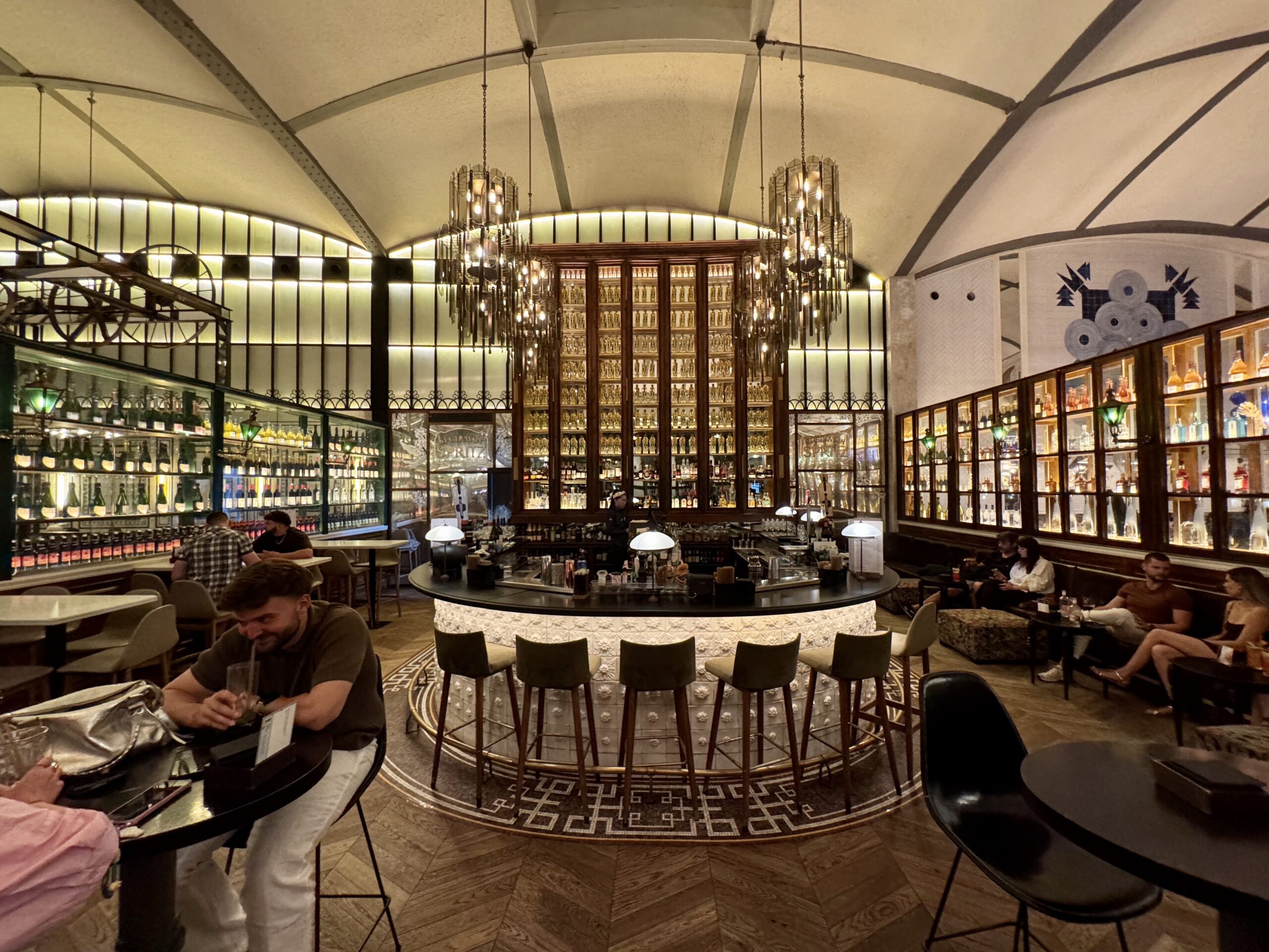 Elegant bar interior with a circular central counter, glass shelves of bottles, and warm hanging chandeliers as patrons sit at surrounding tables.