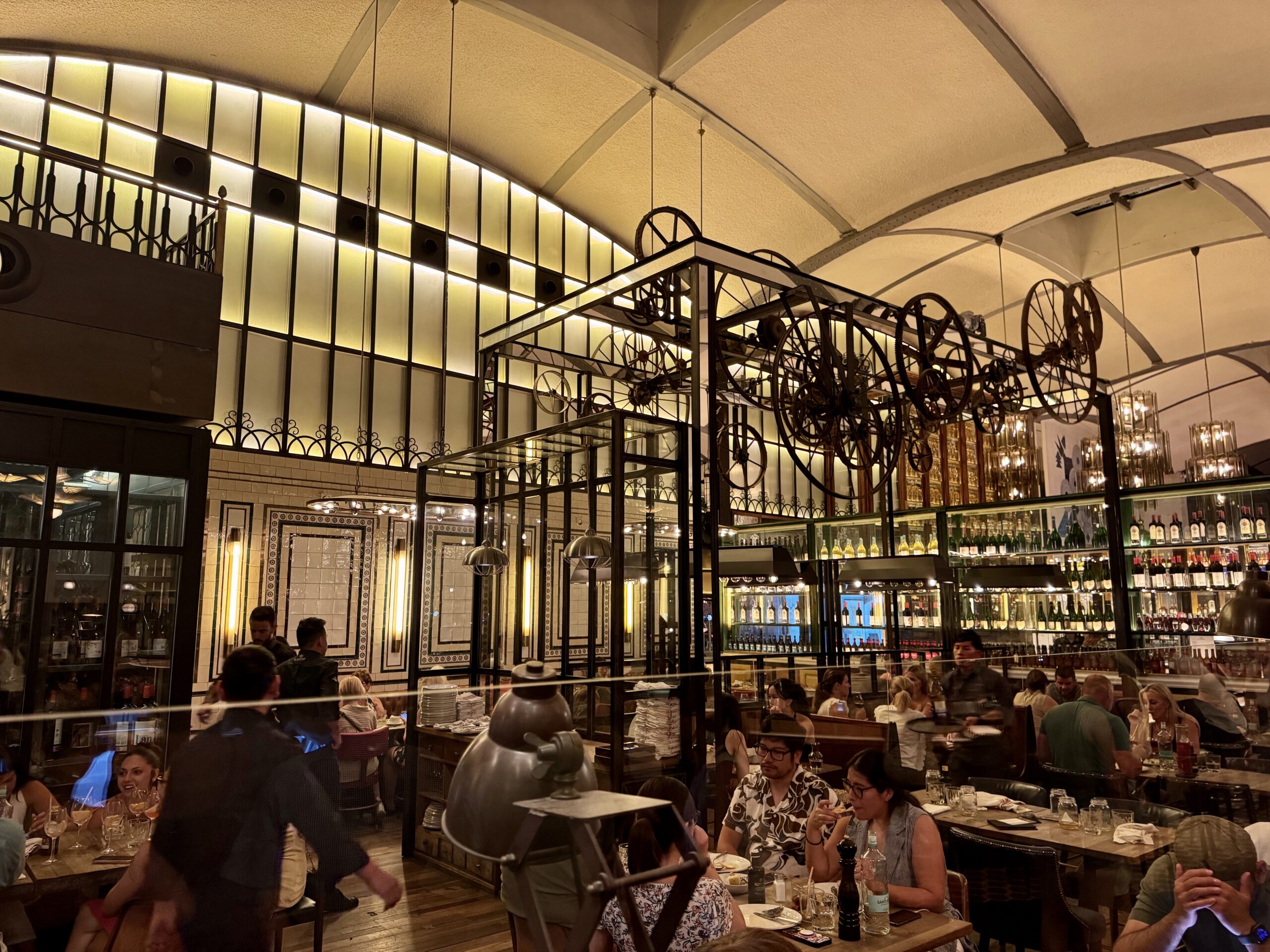 Bustling restaurant interior with diners at tables, glass walls, and vintage machinery decor on display shelves.
