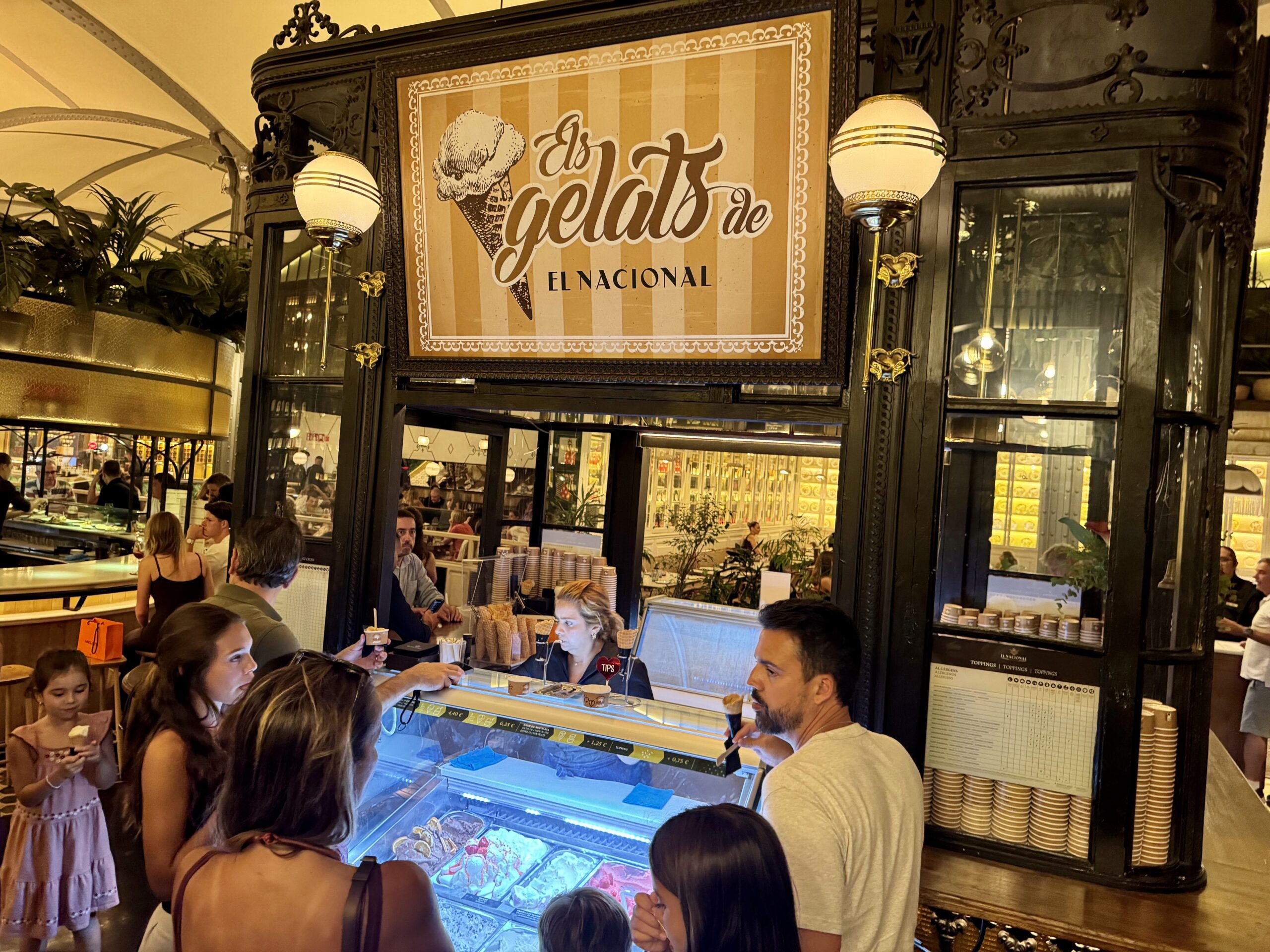 Busy gelato counter inside a vintage-style cafe with customers waiting to order ice cream from a glass display case in front of them.