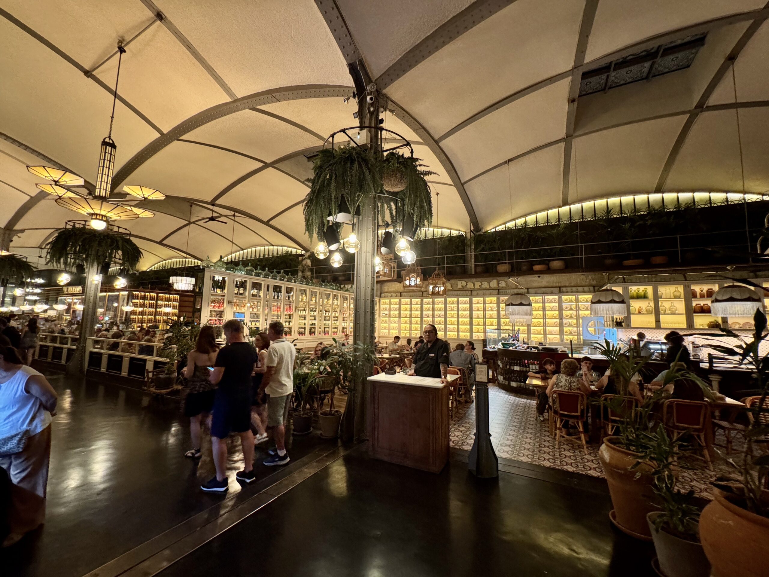 Indoor market dining area with arched metal beams, hanging ferns, and warm pendant lighting as people sit at tables and browse displays.