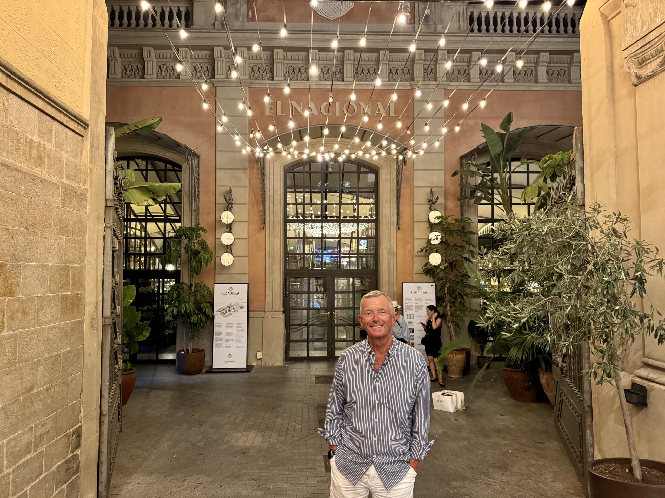 Smiling man in a striped shirt stands in a warmly lit courtyard with string lights and potted plants.