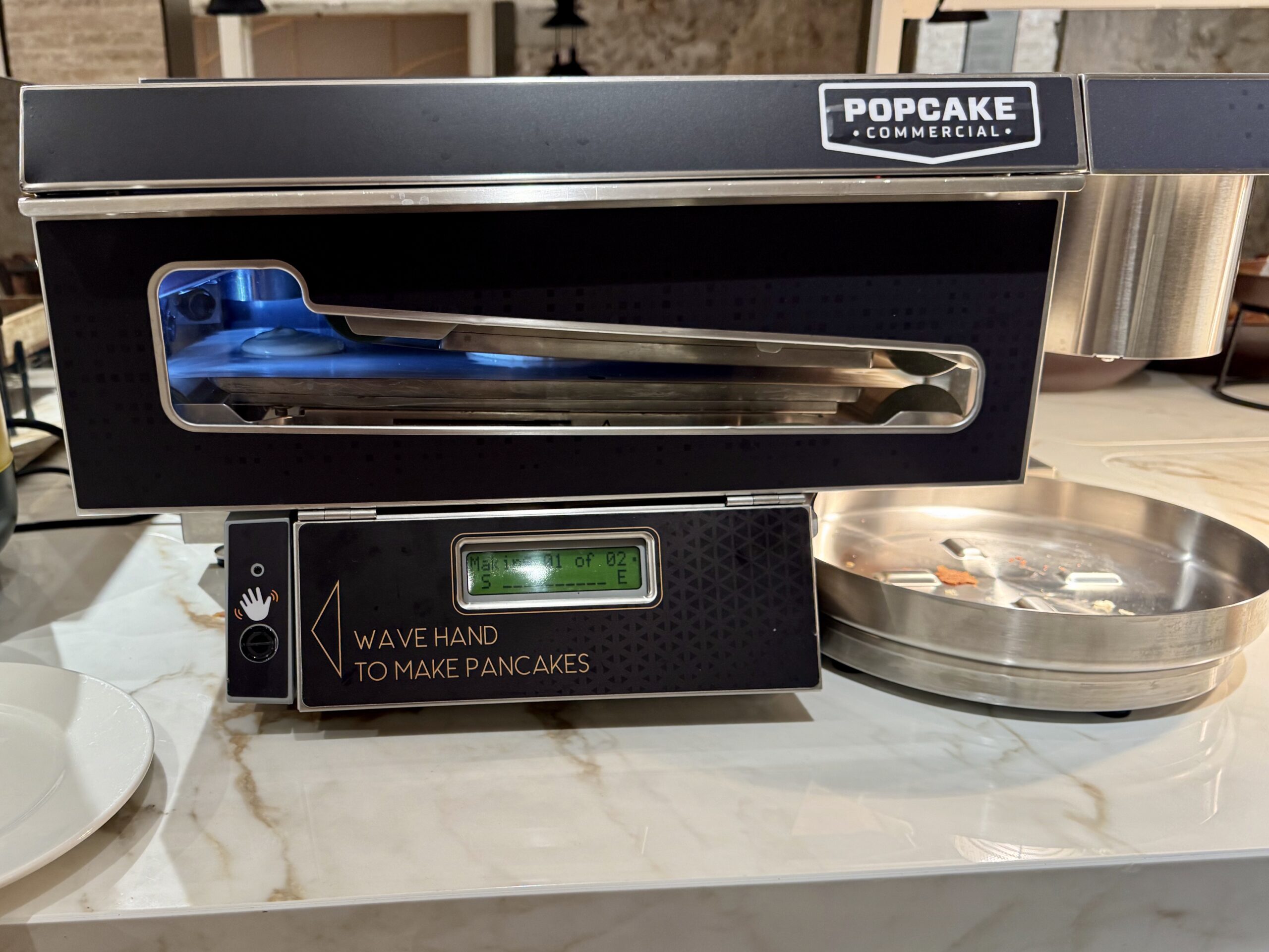 Commercial pancake vending/maker machine labeled 'WAVE HAND TO MAKE PANCAKES' with blue interior lighting and a touchscreen display below the slot.