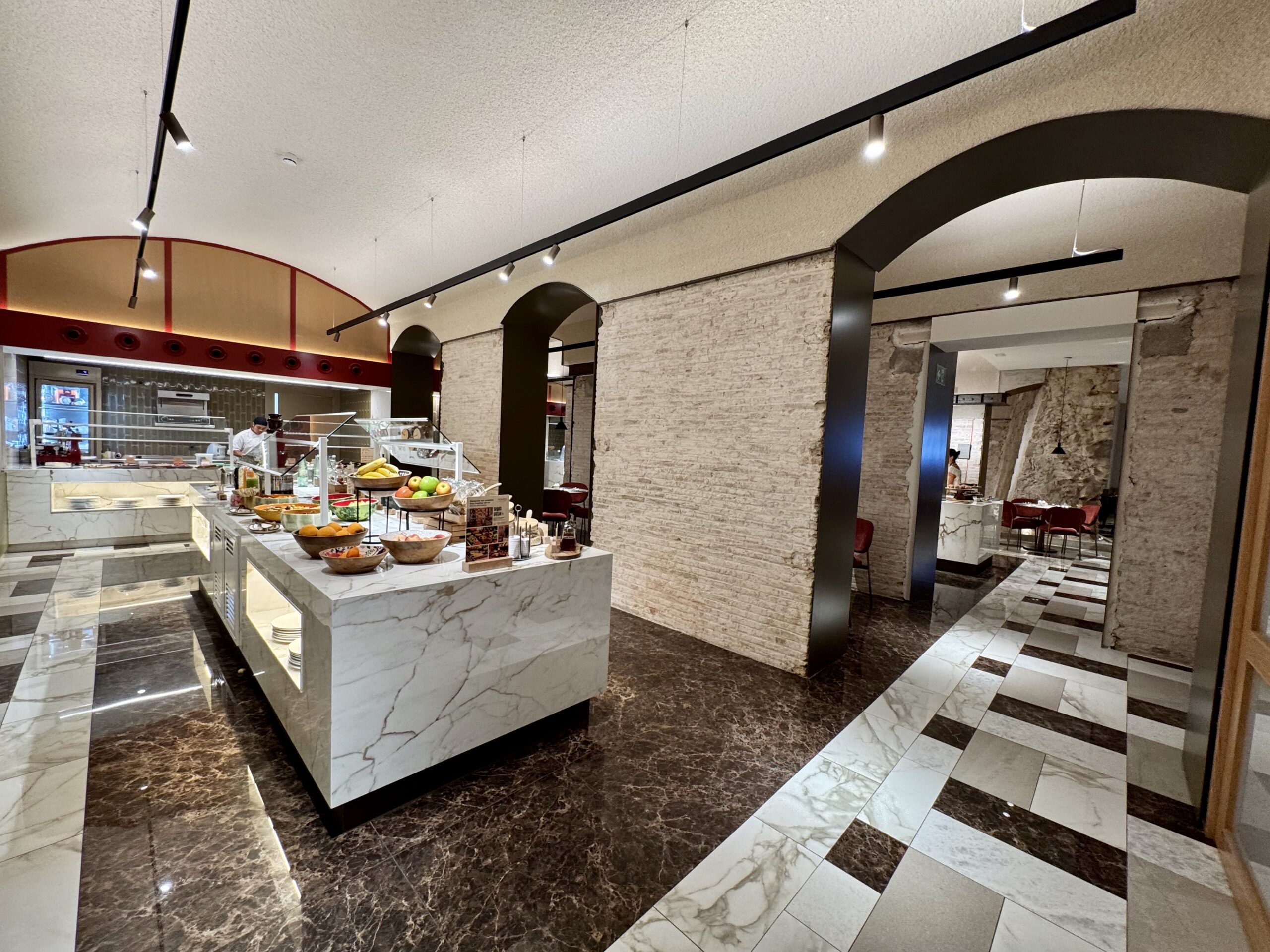 Restaurant dining area with marble fruit counter, warm lighting, and exposed brick walls; arched doorways and track lighting create a modern, upscale atmosphere.