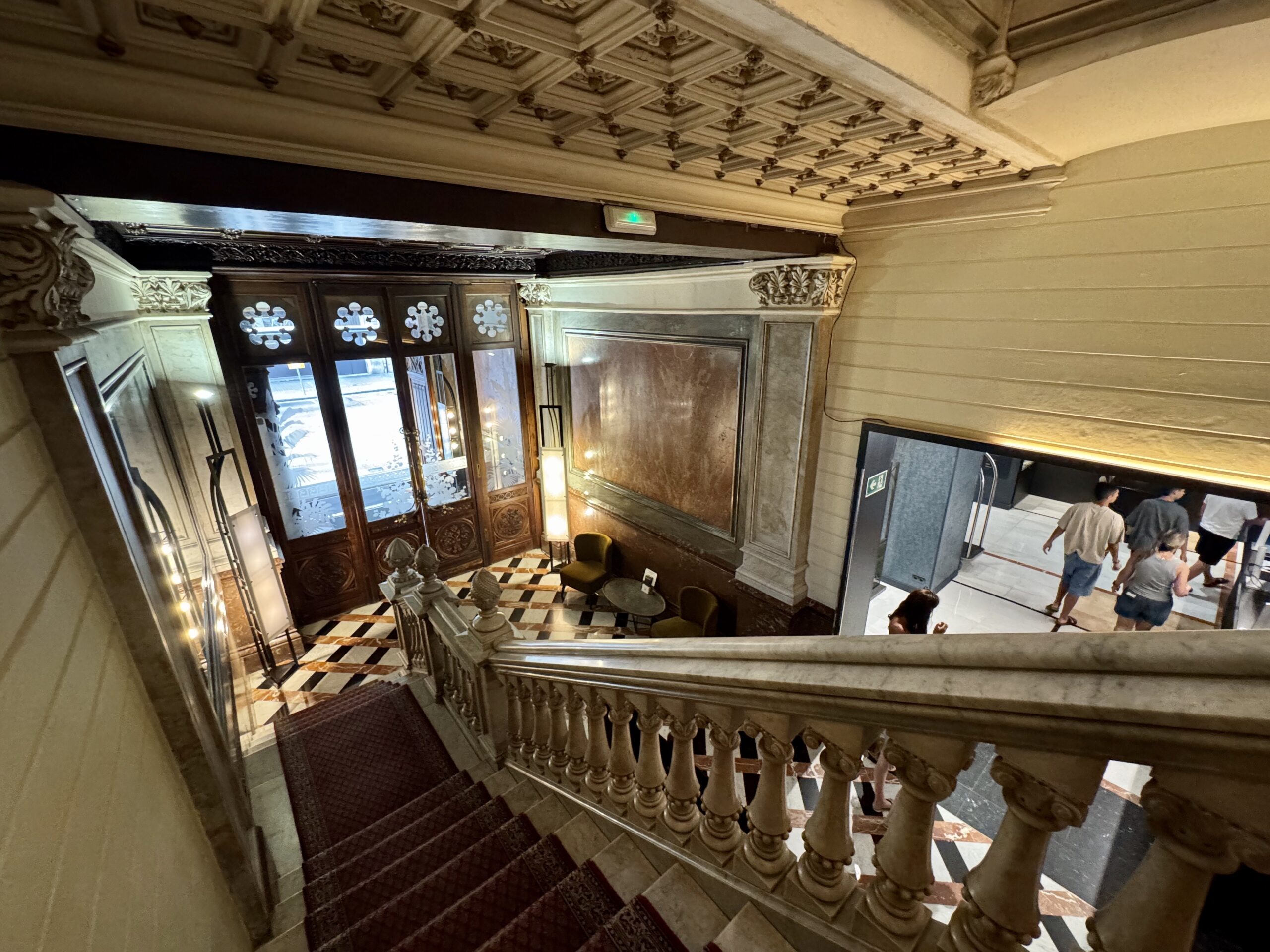 Ornate historic stairwell with carved balustrades, checkered marble floor, and a doorway with decorative panels; people stand near the entrance.