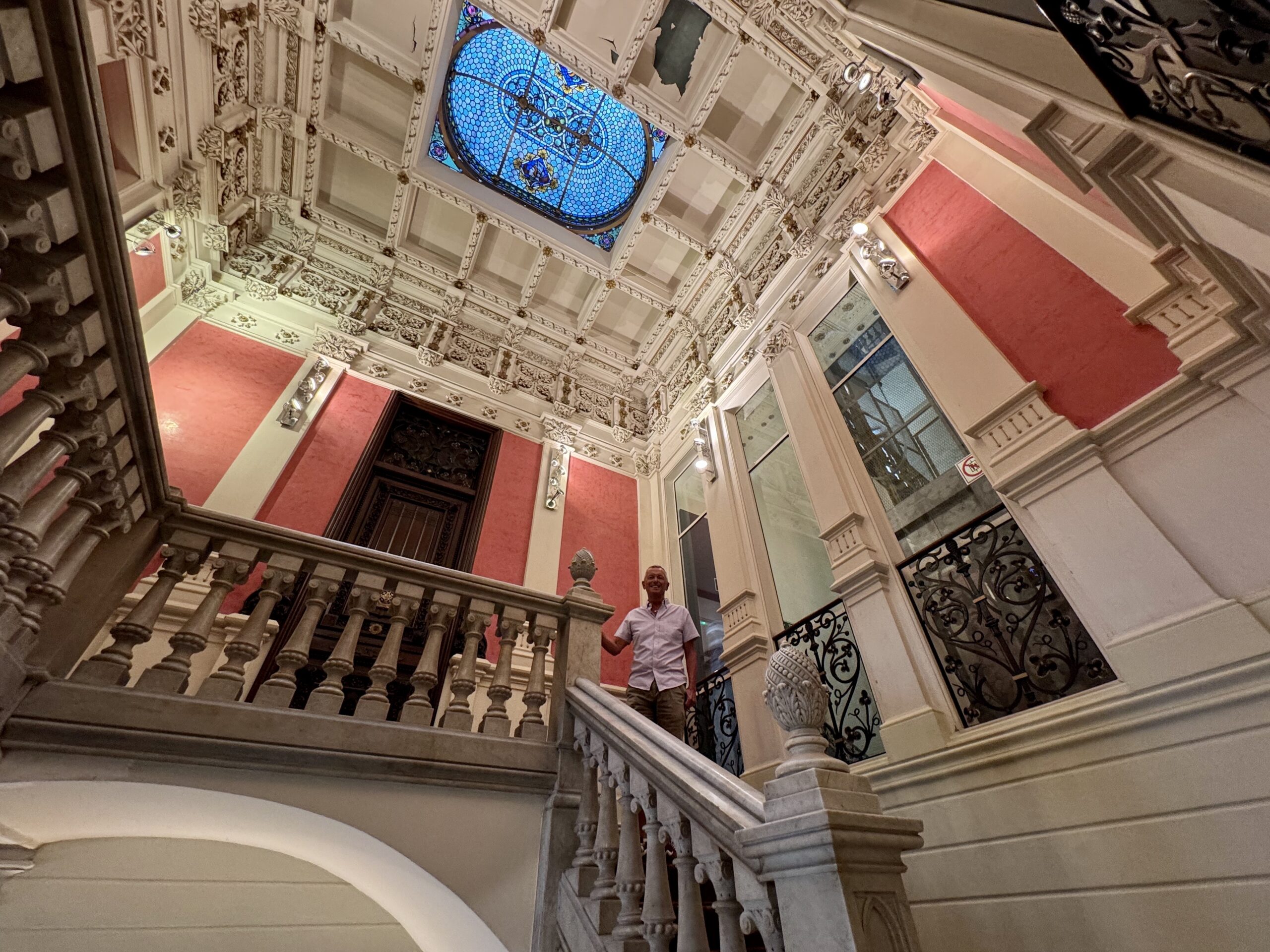 Ornate historic staircase interior with white plaster decoration, marble steps, and a blue stained-glass skylight overhead.