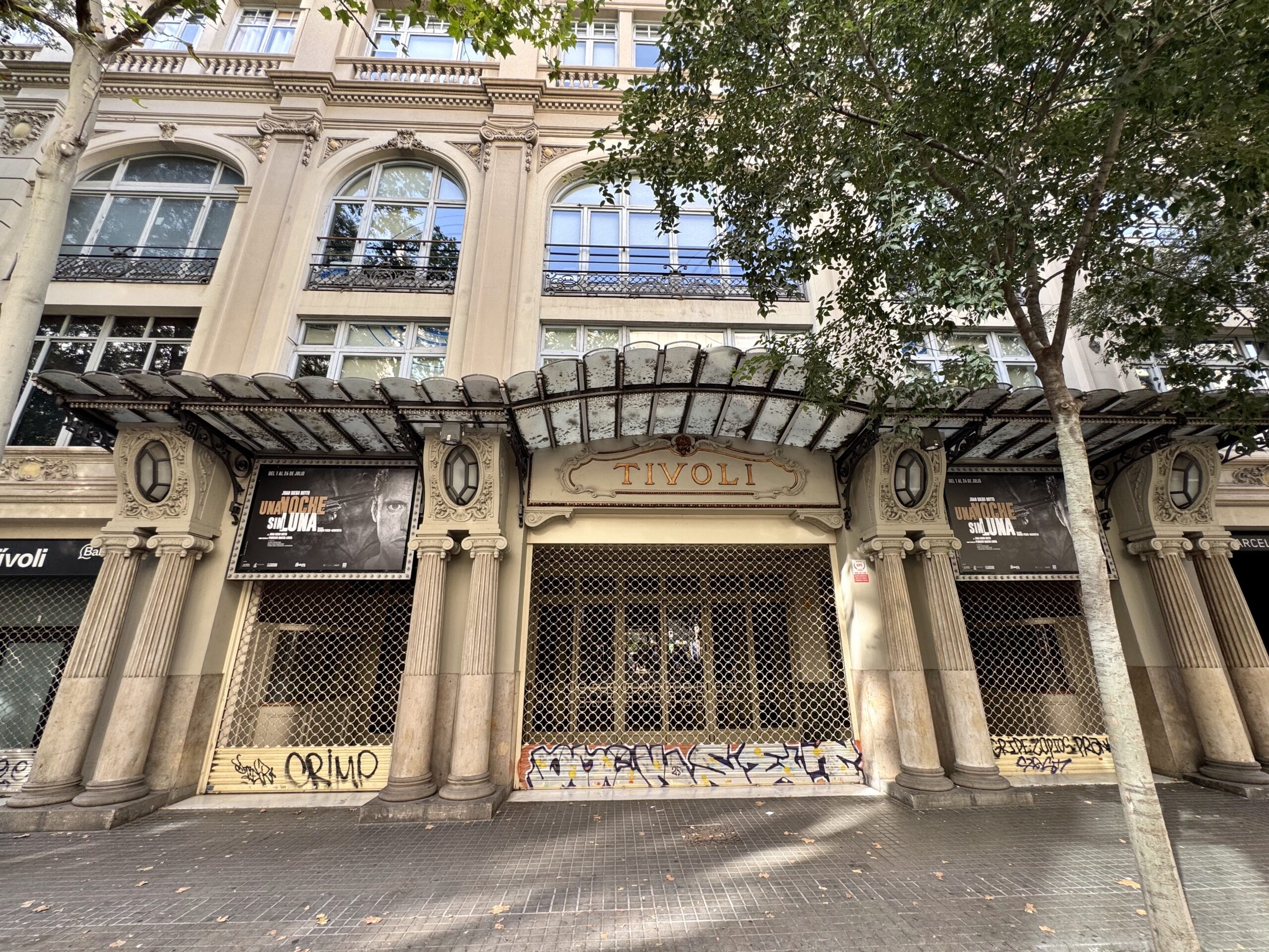 Historic beige building with arched windows and decorative stonework, a leafy tree in front, and graffiti on the shuttered storefronts.