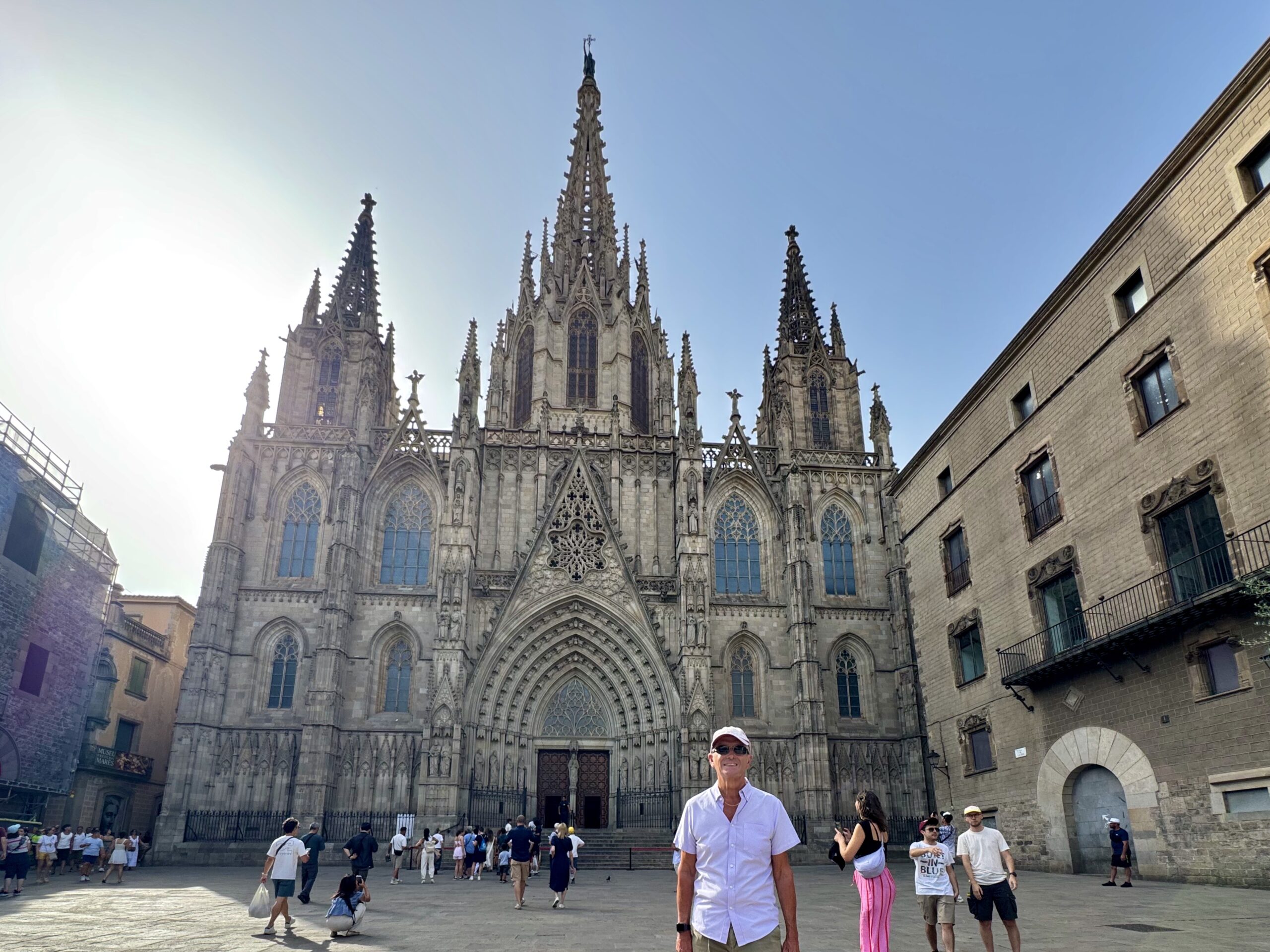 Front view of a Gothic cathedral with ornate spires and a large arched entrance, set in a sunny plaza with tourists nearby.
