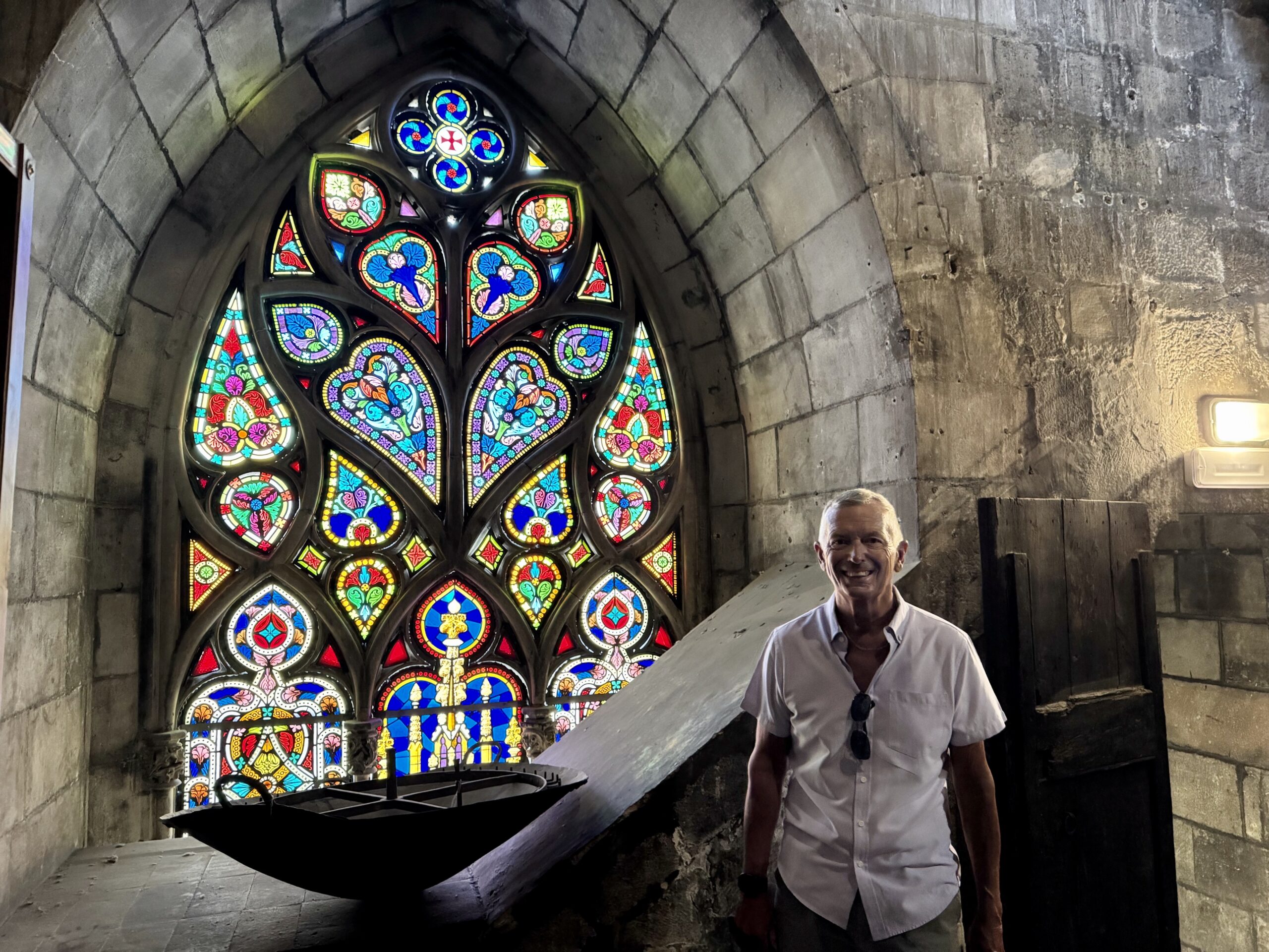 Smiling man in a white shirt stands inside a stone church beside a large colorful stained-glass window.