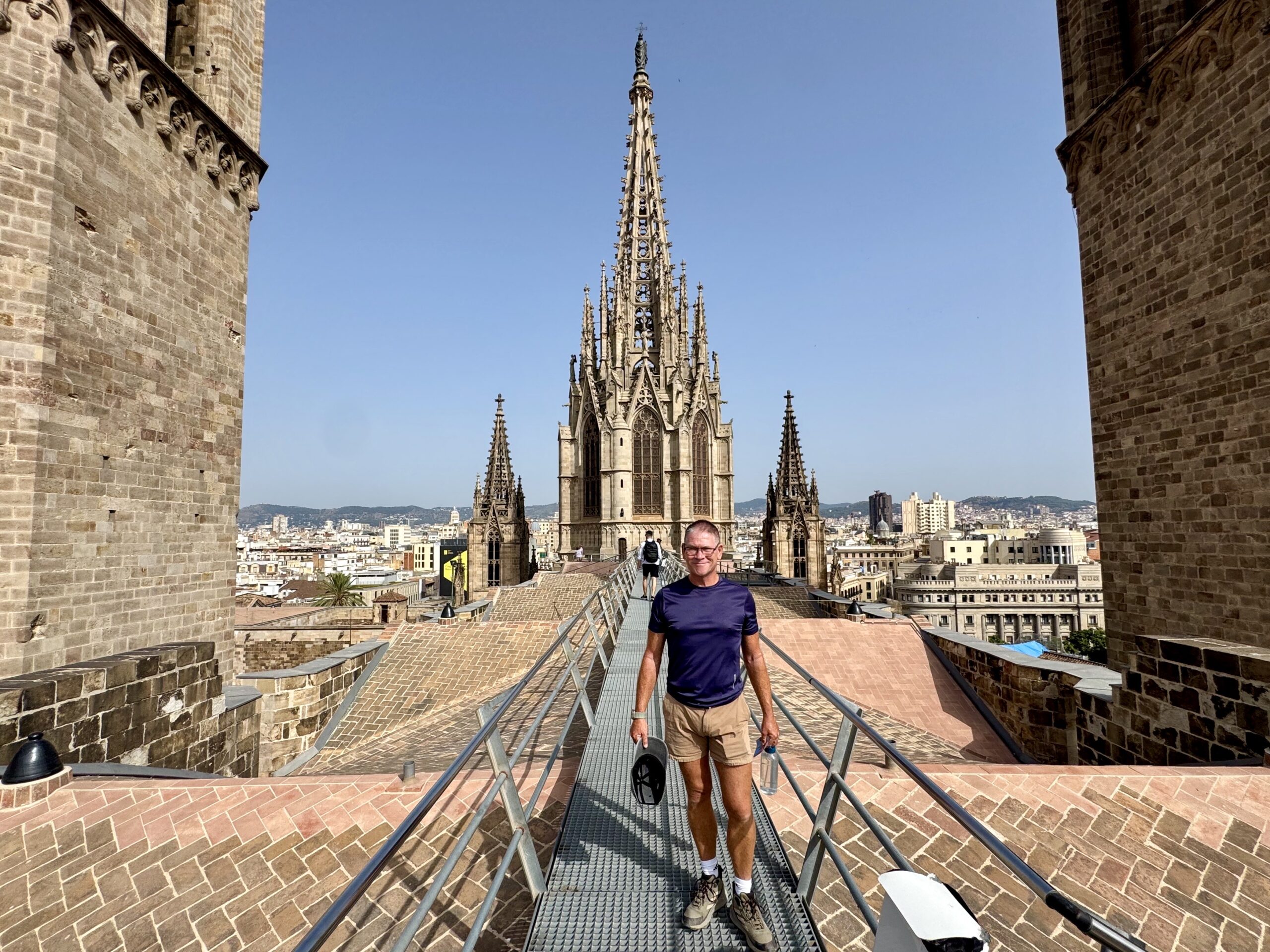 Man walking on a metal walkway atop a historic stone cathedral, with ornate spires in the background and a cityscape beyond.