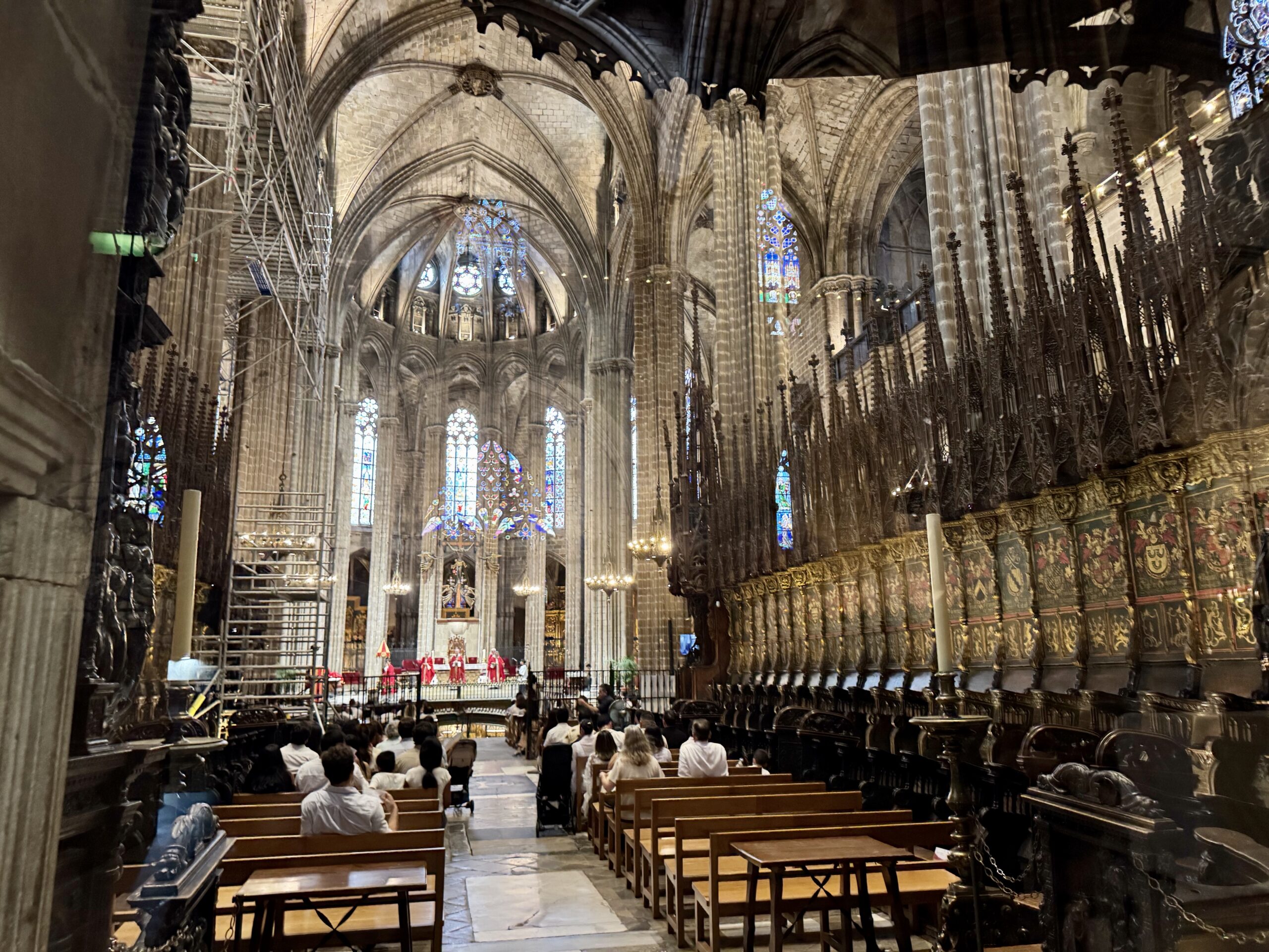 Gothic cathedral nave with tall arches and stained-glass windows, ornate railings, and scaffolding on the left; people seated in pews.