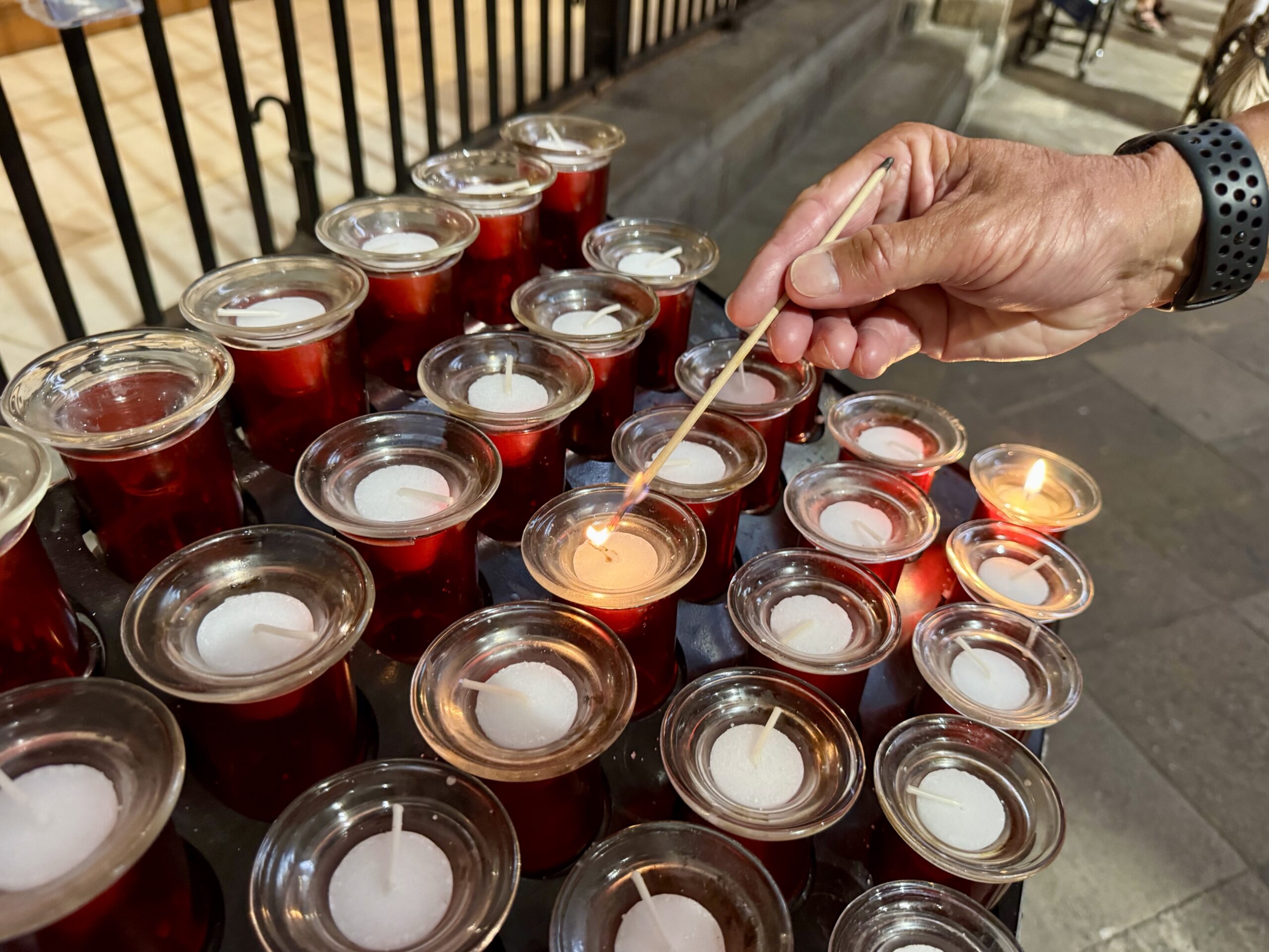 A hand with a perforated wristband uses a wooden skewer to light or adjust red votive candles arranged on a surface.