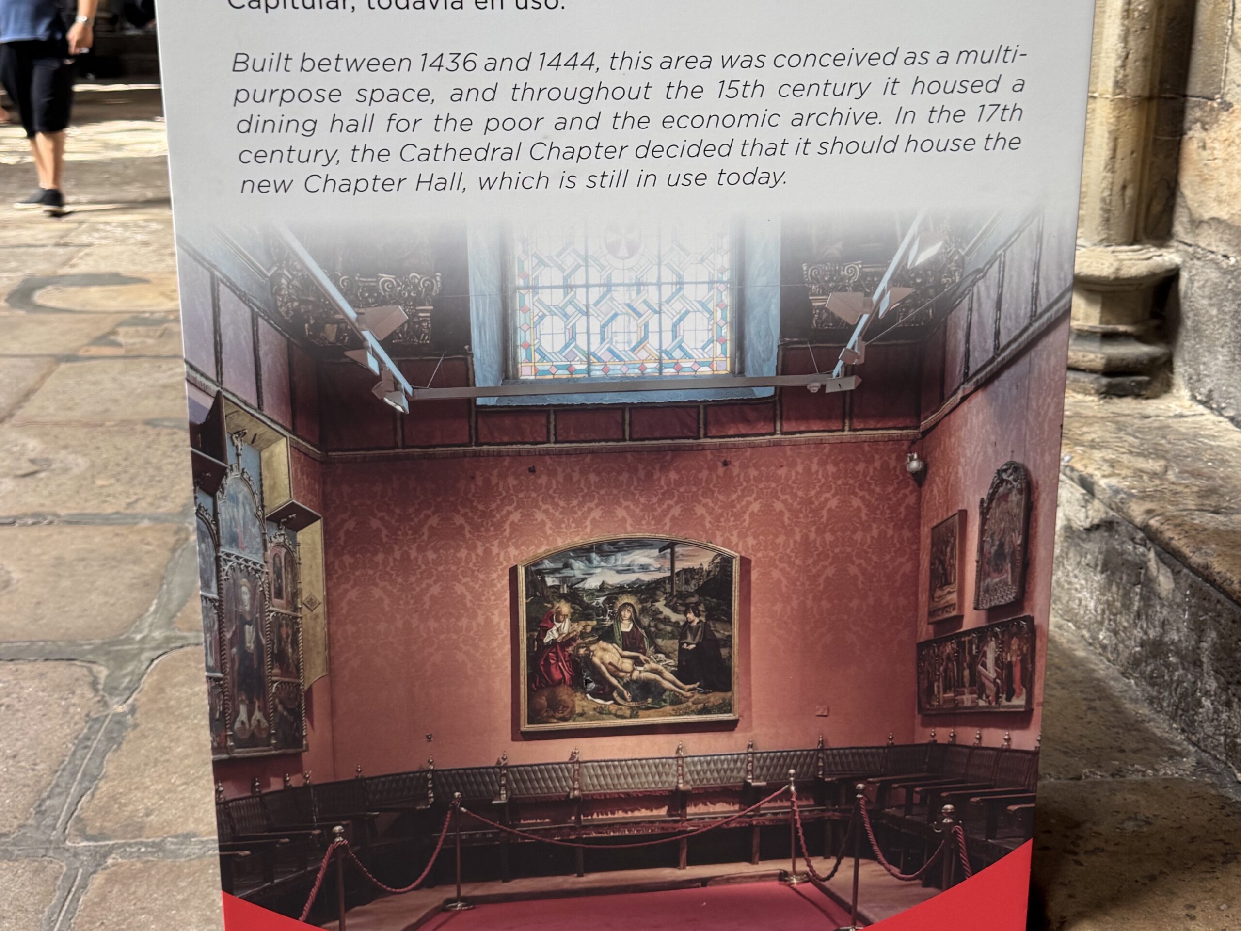 Exhibition panel showing classical paintings on red patterned walls with rope barriers in front.