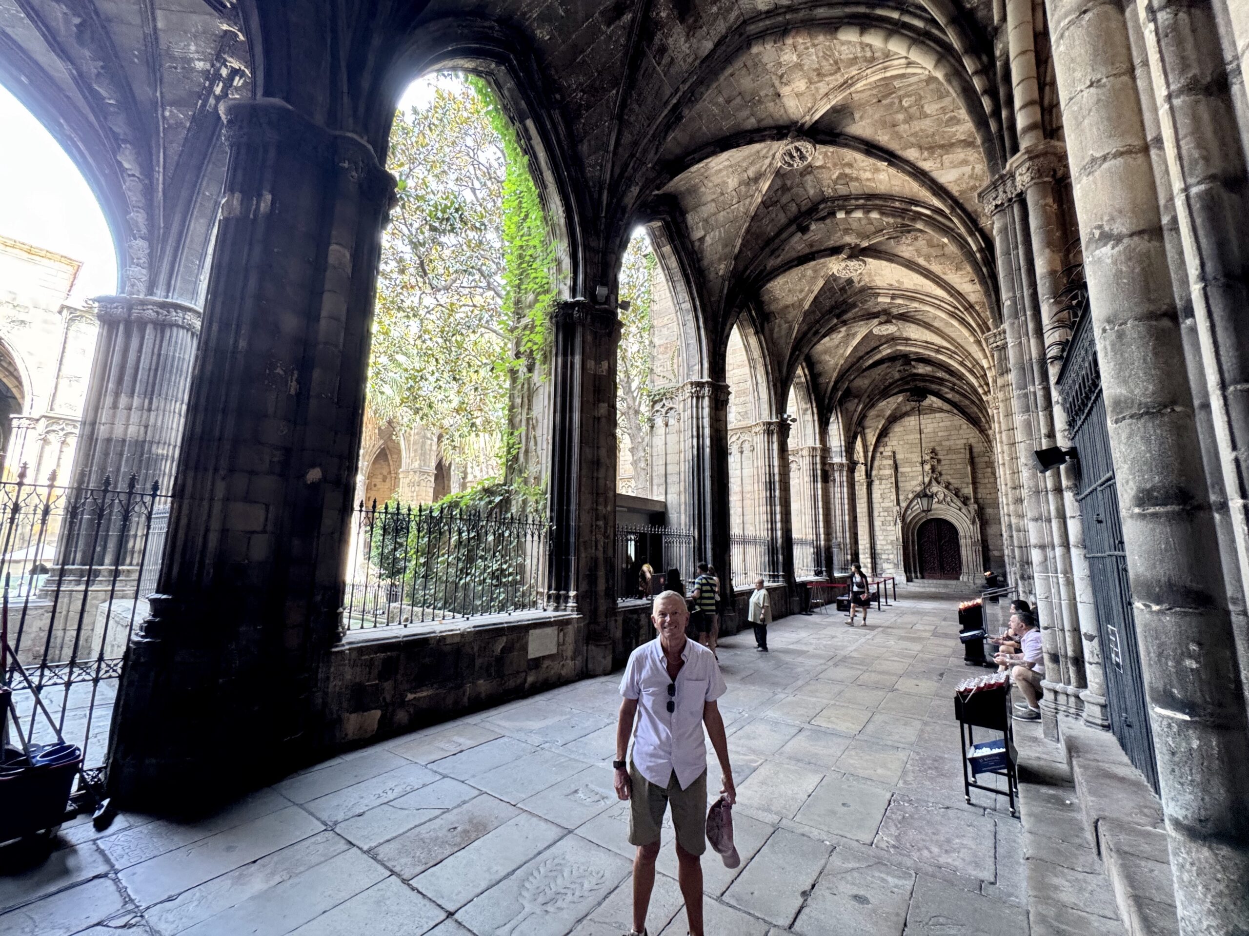 Man in a white shirt and khaki shorts stands in a historic stone cloister with tall arches behind him.