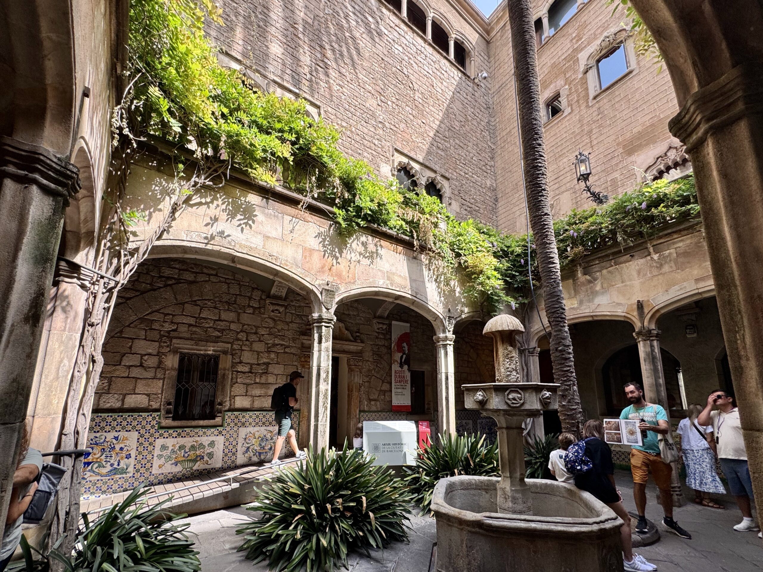Historic stone courtyard with arches, vines, and a tall palm tree; visitors examine information boards and displays.