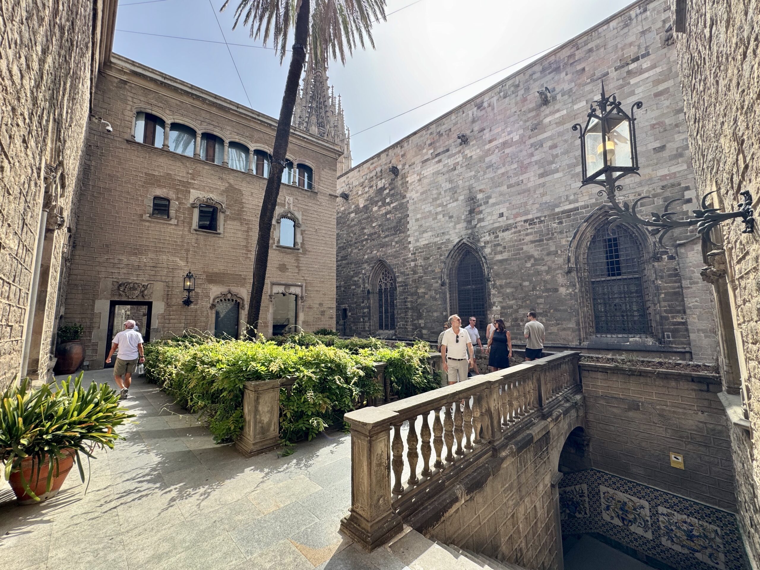 Stone courtyard with Gothic arched windows, palm tree, and ornate lamp; people walk and chat in warm daylight.