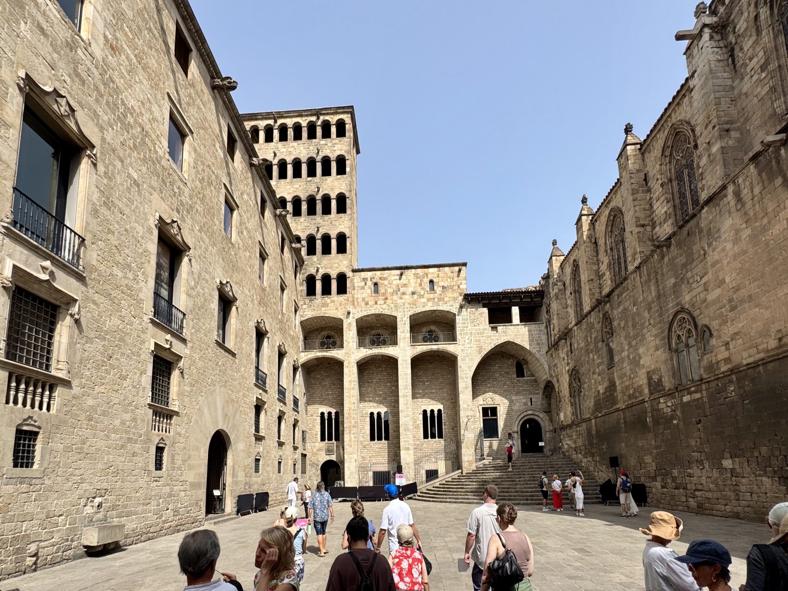 Stone courtyard surrounded by beige stone buildings with arched arcades; tourists walk up and down steps toward a central doorway.