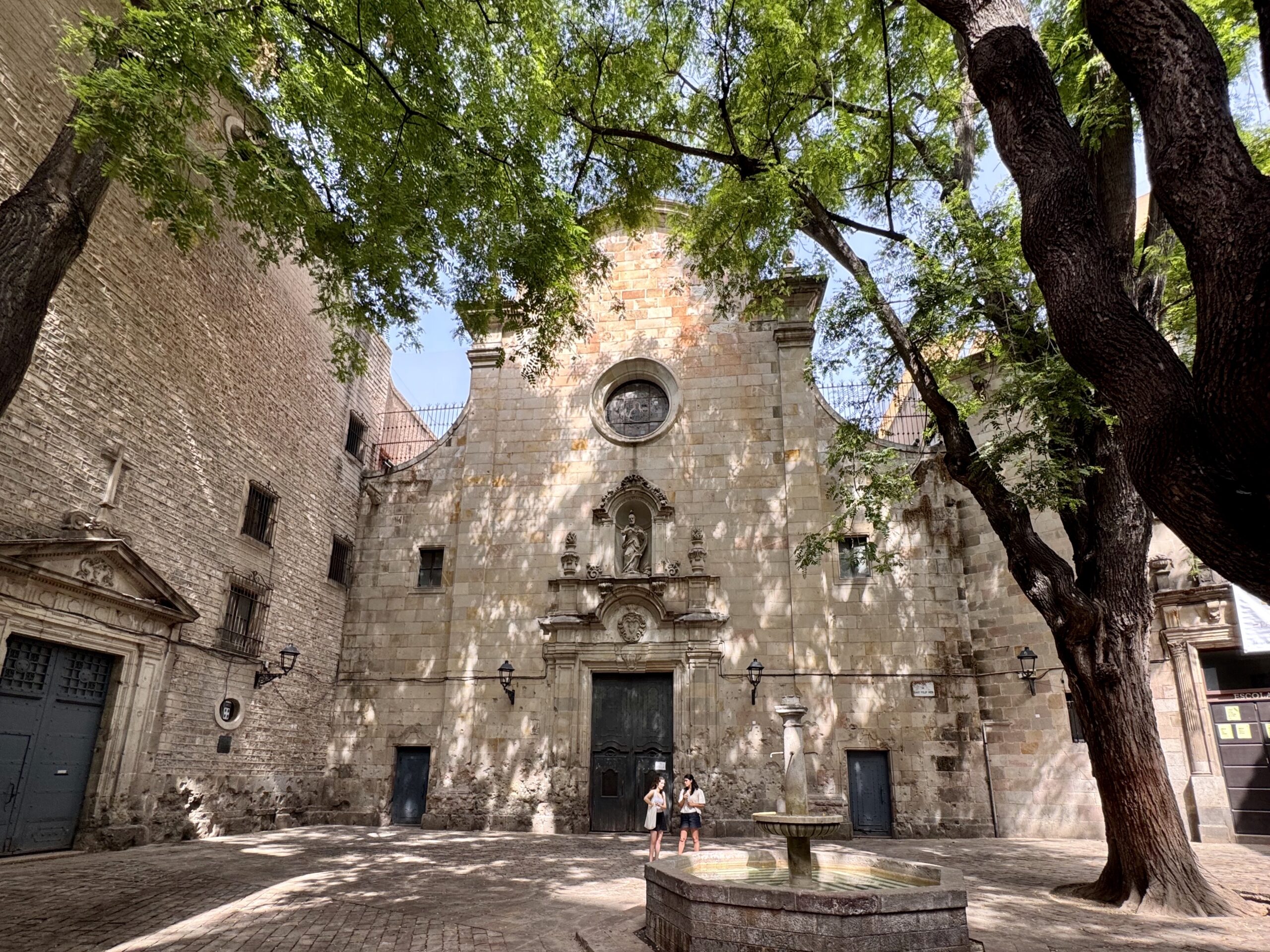 Historic stone courtyard with arched doorway, circular window, and statue reliefs, shaded by lush green trees.