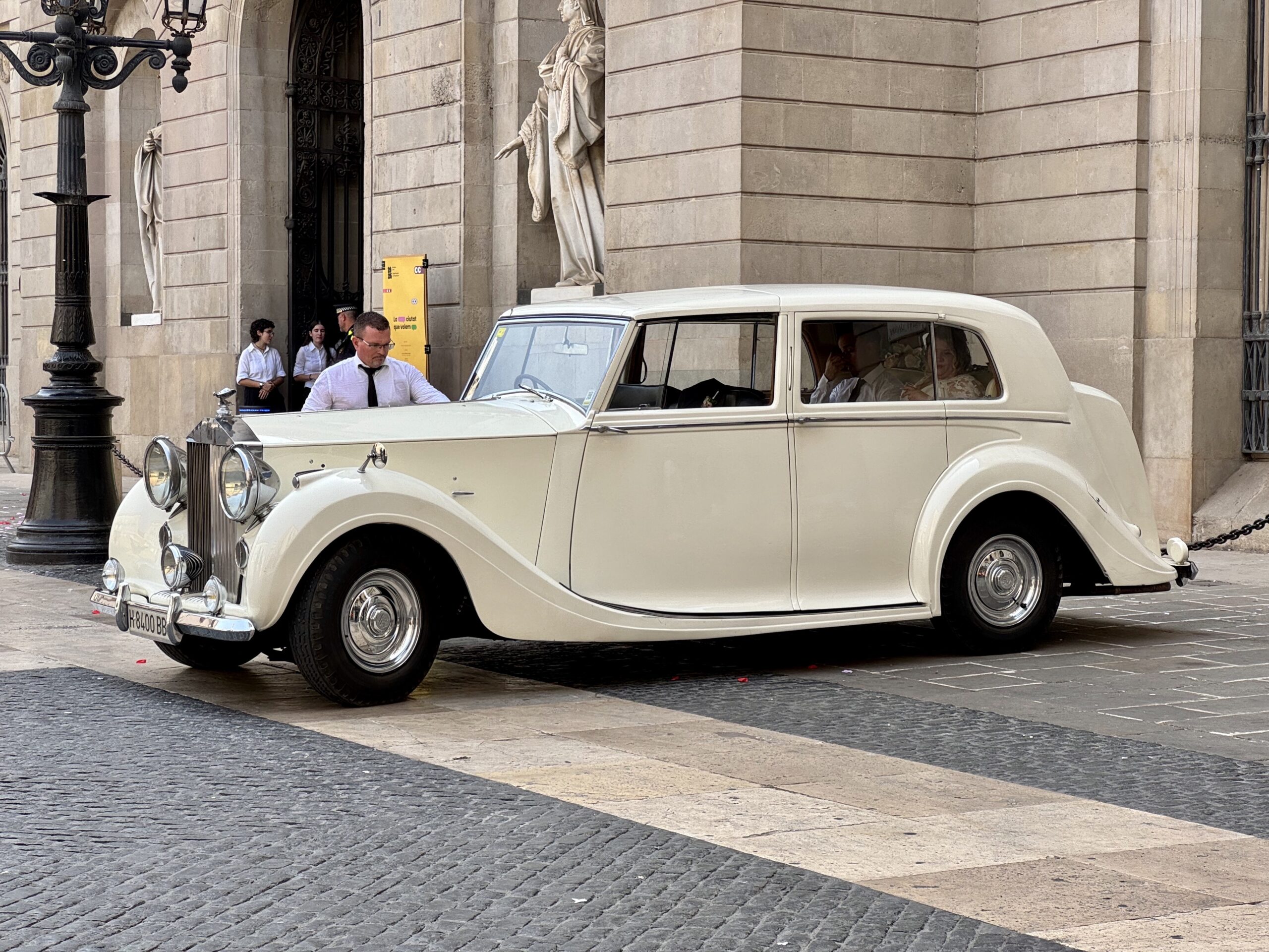 White vintage car parked on cobblestone street in front of a stone building with statues; people in white shirts gathered near the entrance.