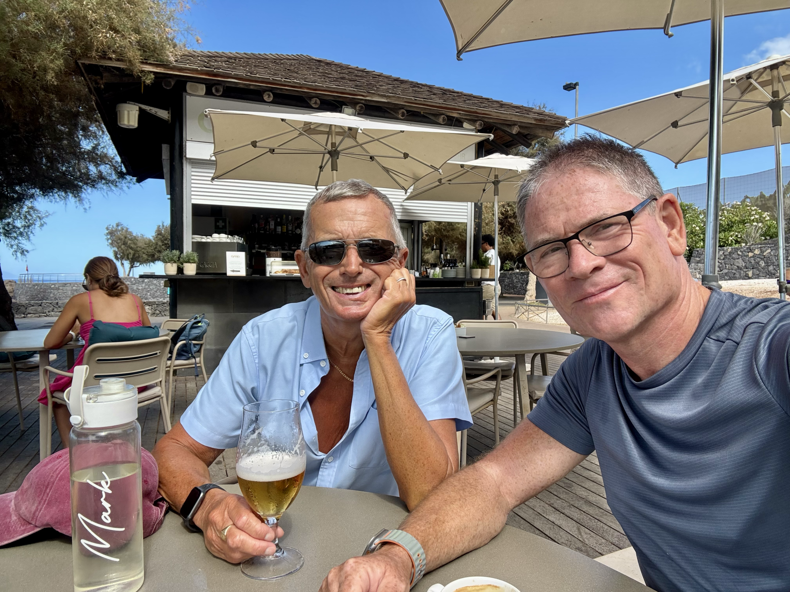 Two men sit at an outdoor cafe table with drinks, smiling at the camera; beer glasses, umbrellas, and a bar in the background.