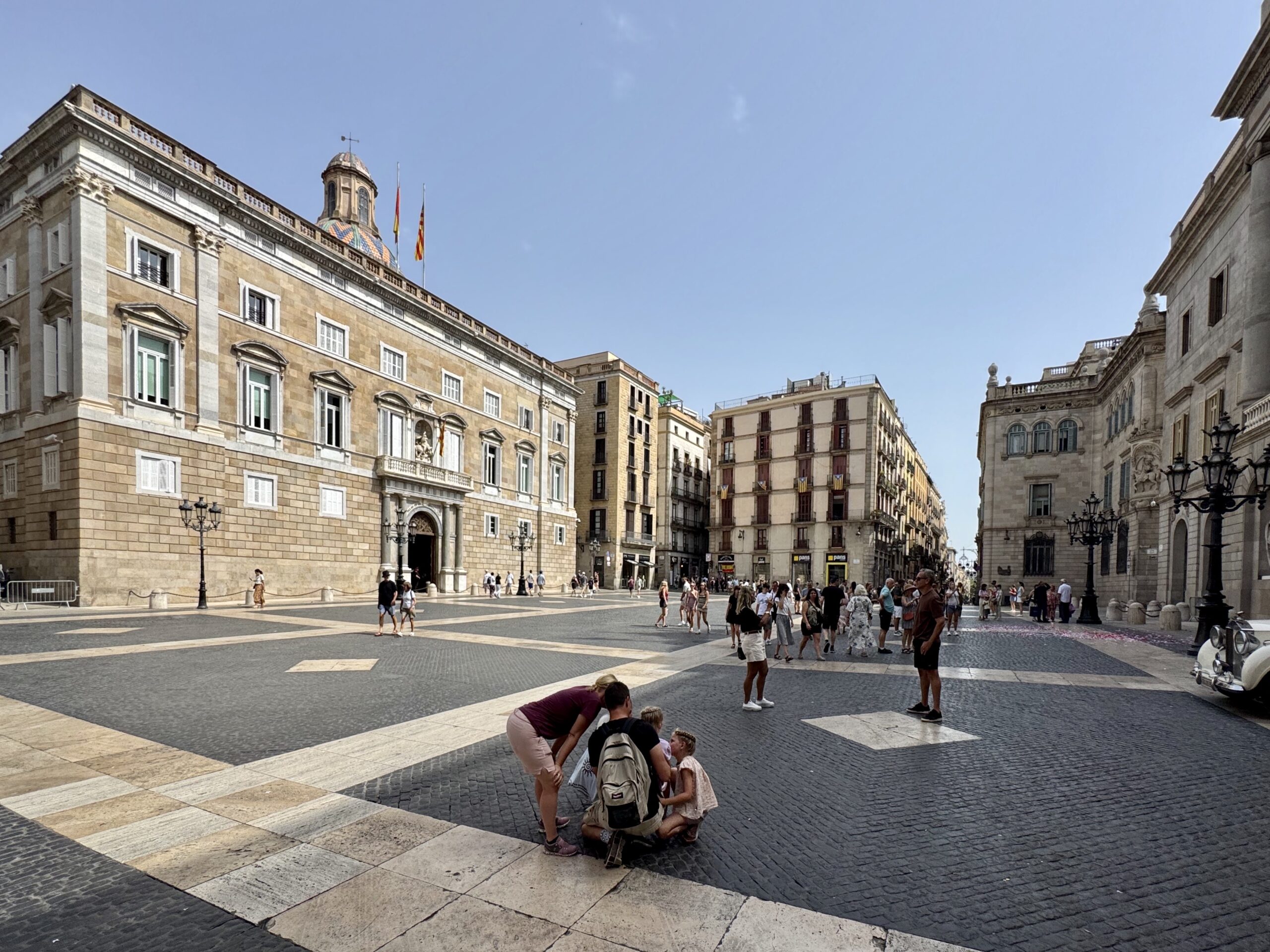 Sunlit European city square with grand stone buildings, a domed tower, and striped pavement; pedestrians walk and chat in the open plaza.
