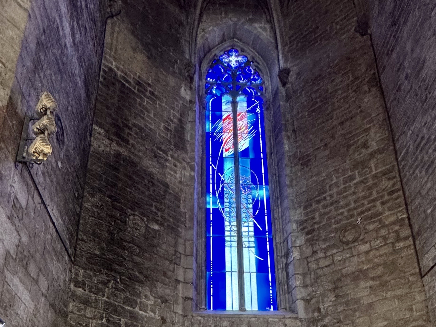 Tall blue stained-glass window in a stone church, with a small carved statue plaque on the left wall.