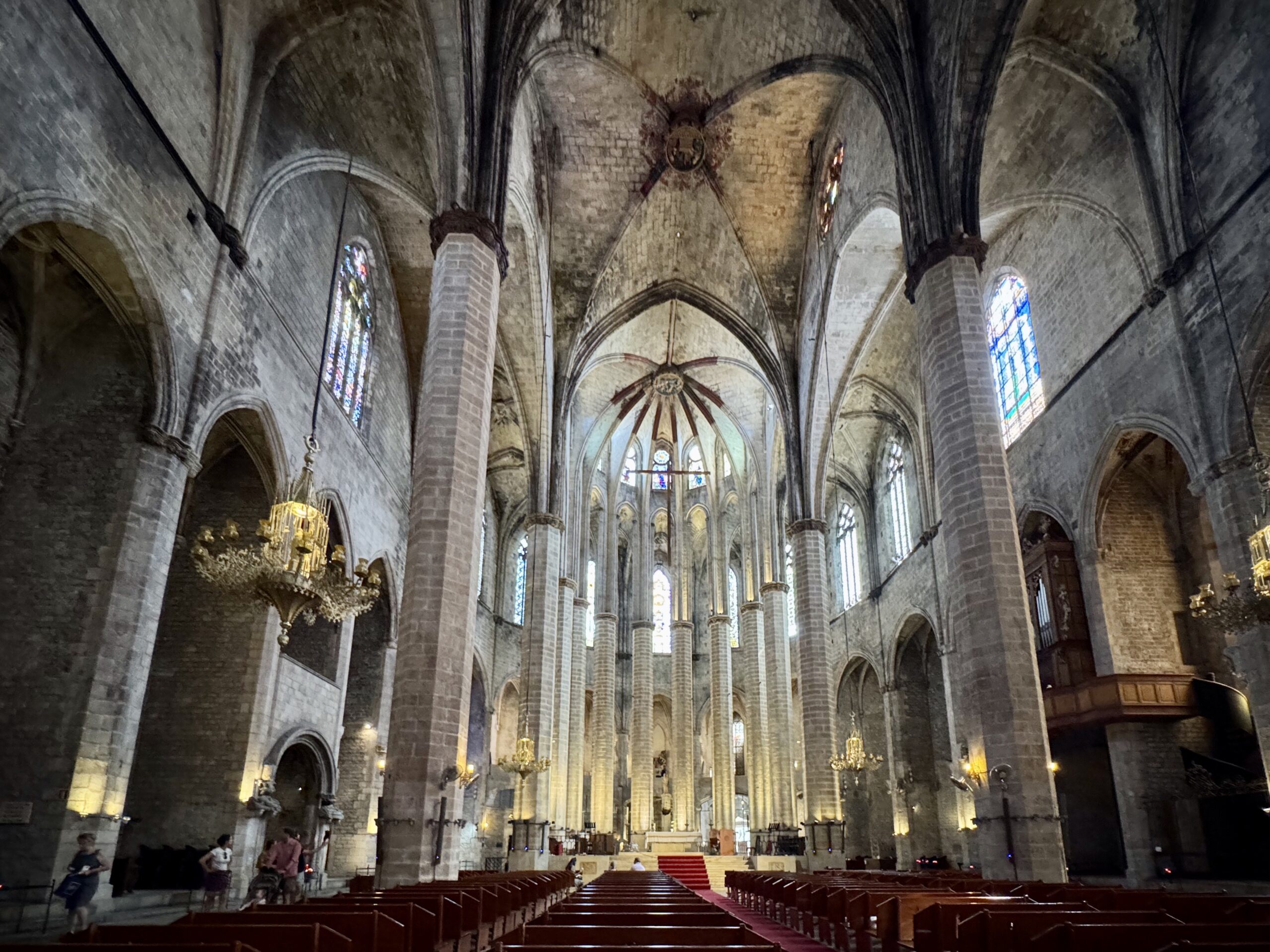 Interior of a Gothic cathedral with tall stone columns, vaulted arches, and stained-glass windows illuminated by warm lights behind the pews.