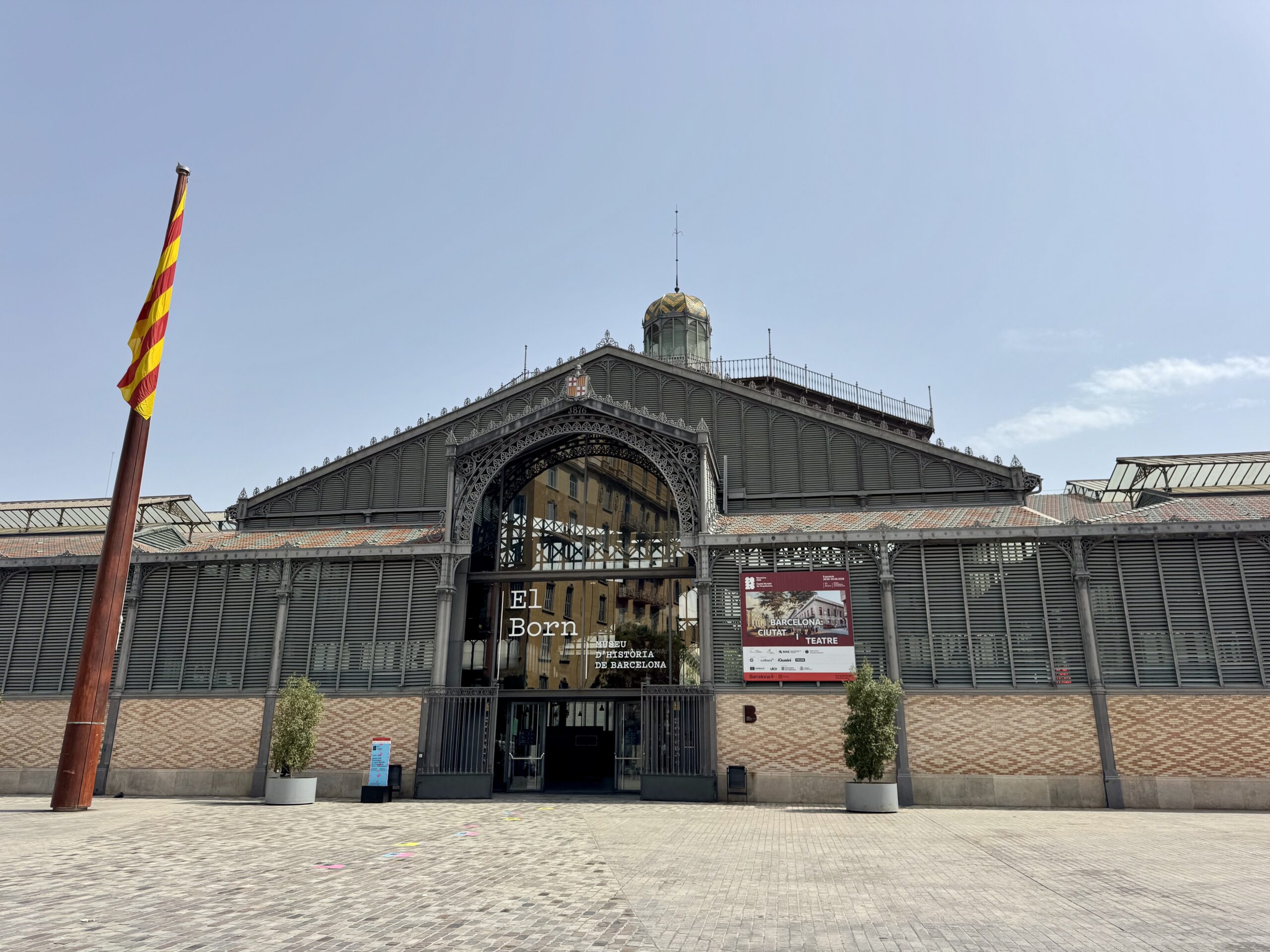 Facade of an ornate historic building with intricate ironwork and arched entrance; a striped Catalan flag on the left against a blue sky.