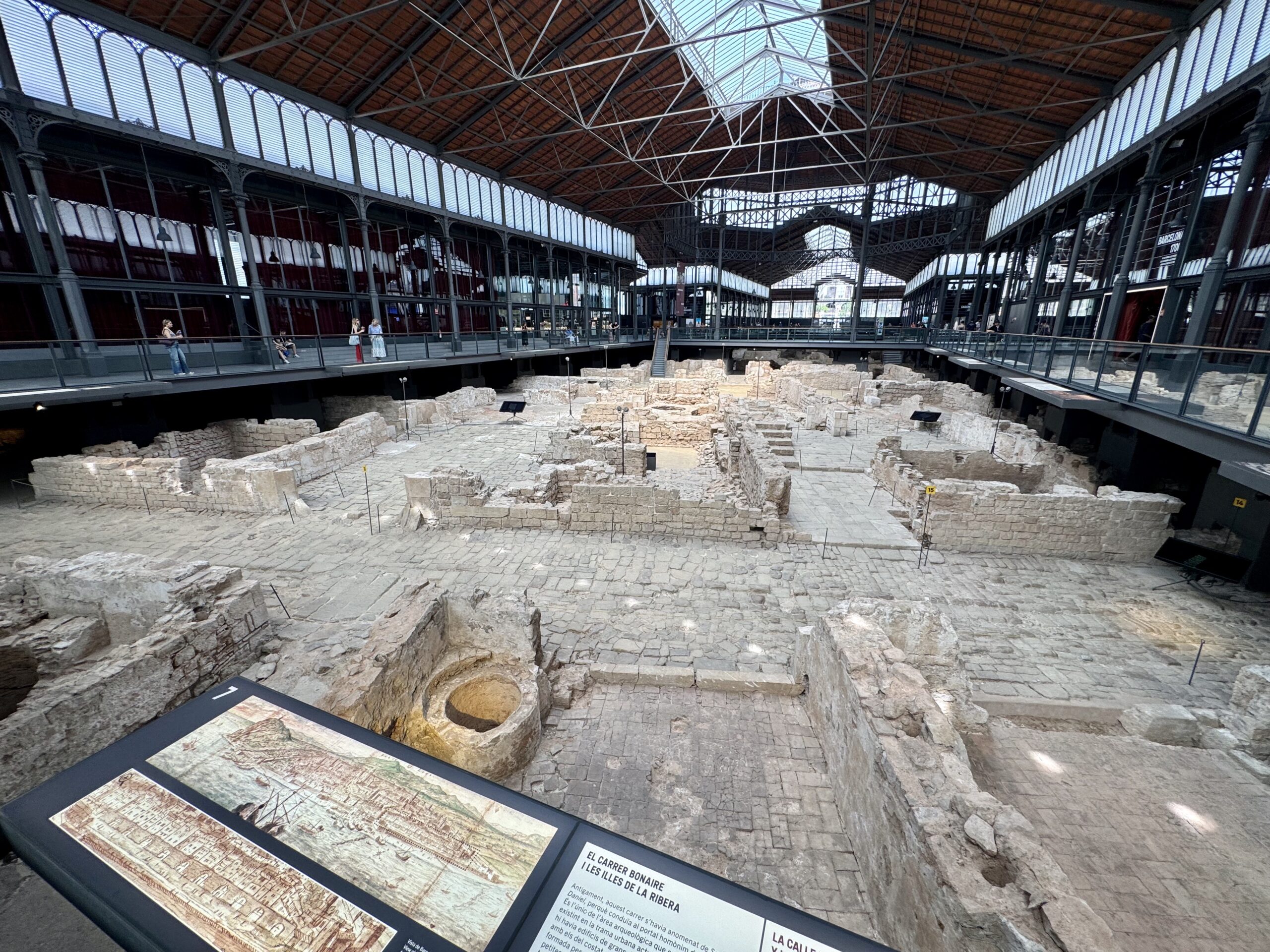 Museum interior over ancient ruins: glass-roofed atrium with metal arches and a walkway around exposed stone walls; visitors observe displays.