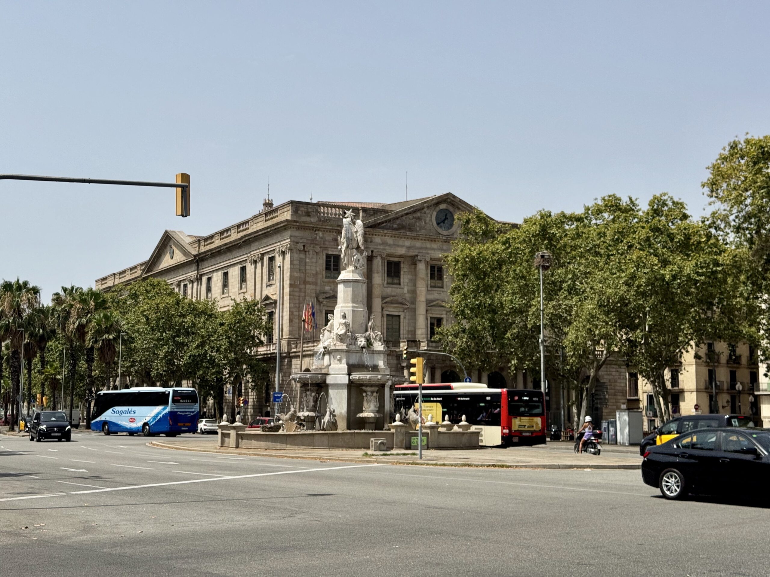 Large neoclassical government building with a commanding statue fountain in front, set in a tree-lined city square under a clear sky