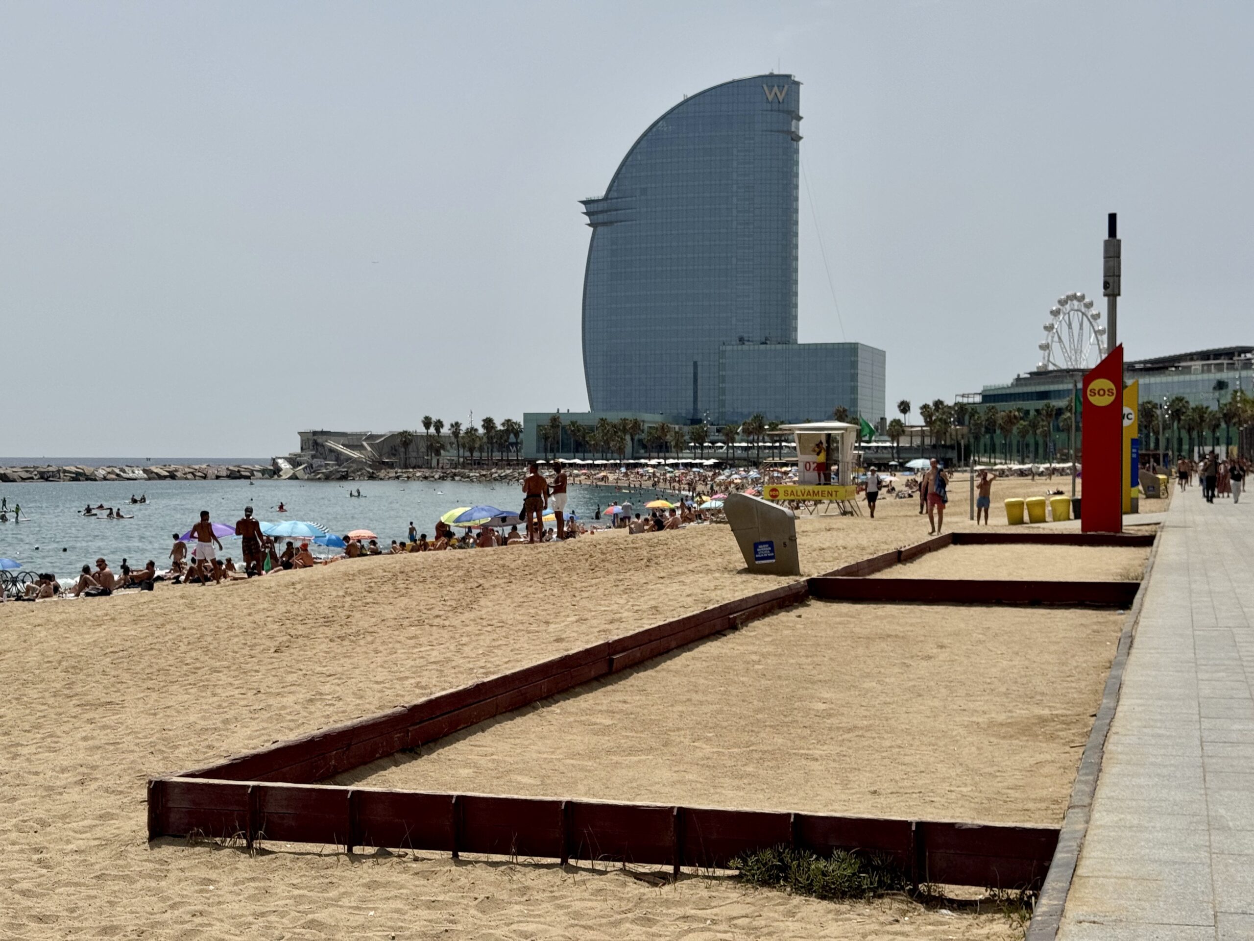 Sail-shaped glass hotel by the beach with sunbathers, umbrellas, and a distant Ferris wheel visible in the background.