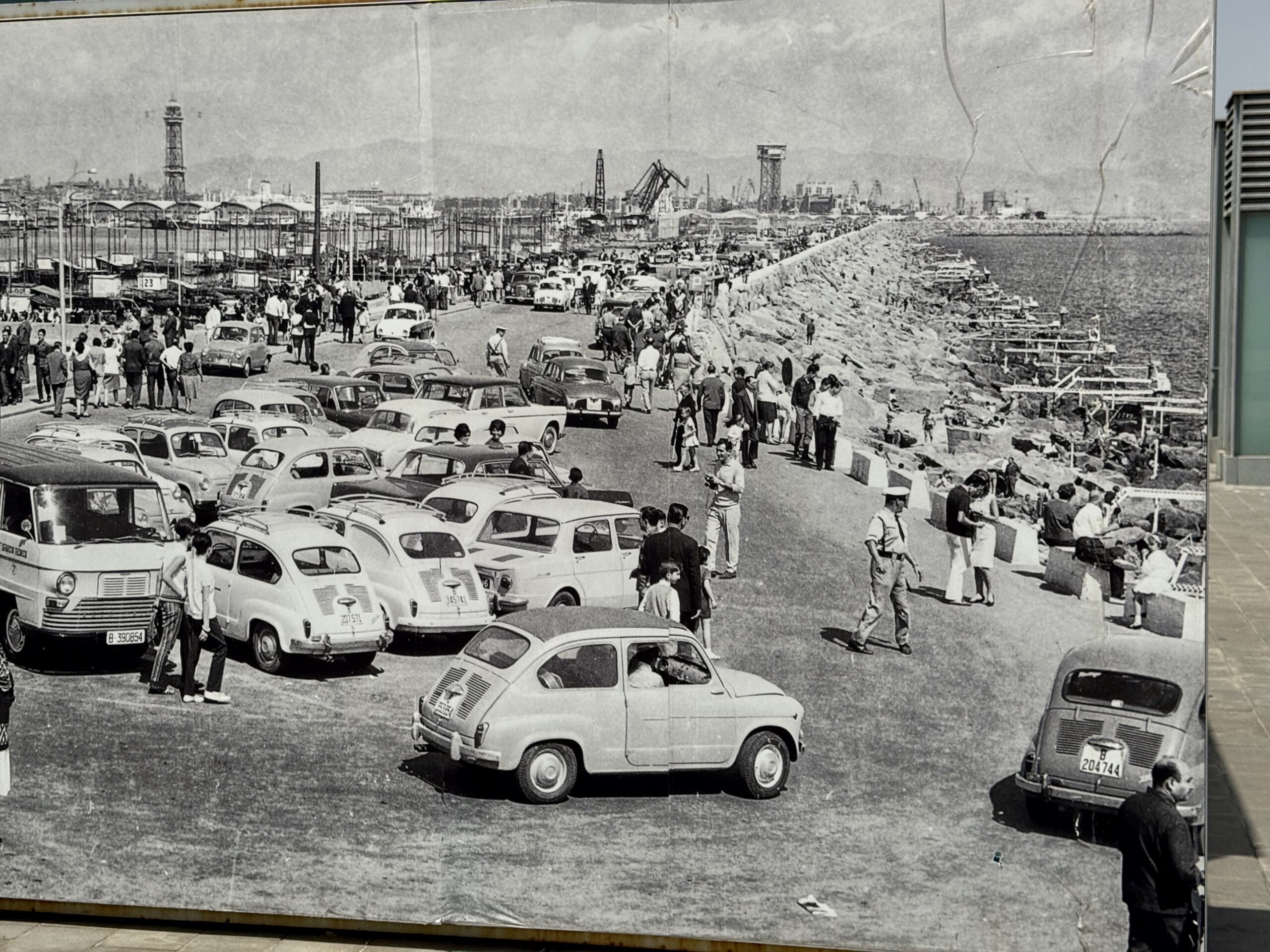 Historic black-and-white photo of a crowded seaside boardwalk, with vintage cars parked along a road and people strolling by the shore; midway background shows amusement structures and a pier.
